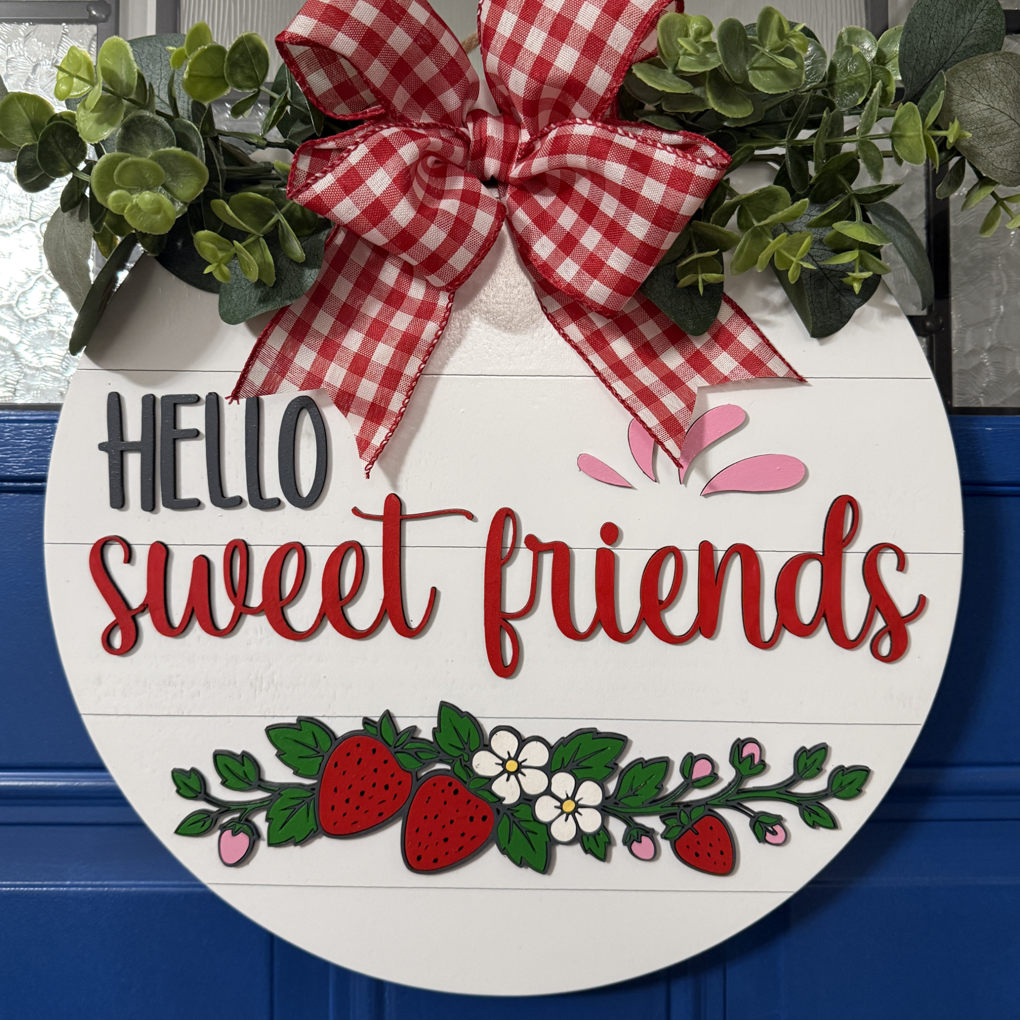 Front view of Hello Sweet Friends strawberry door hanger with raised red lettering, hand-painted strawberries, and red gingham bow on blue door