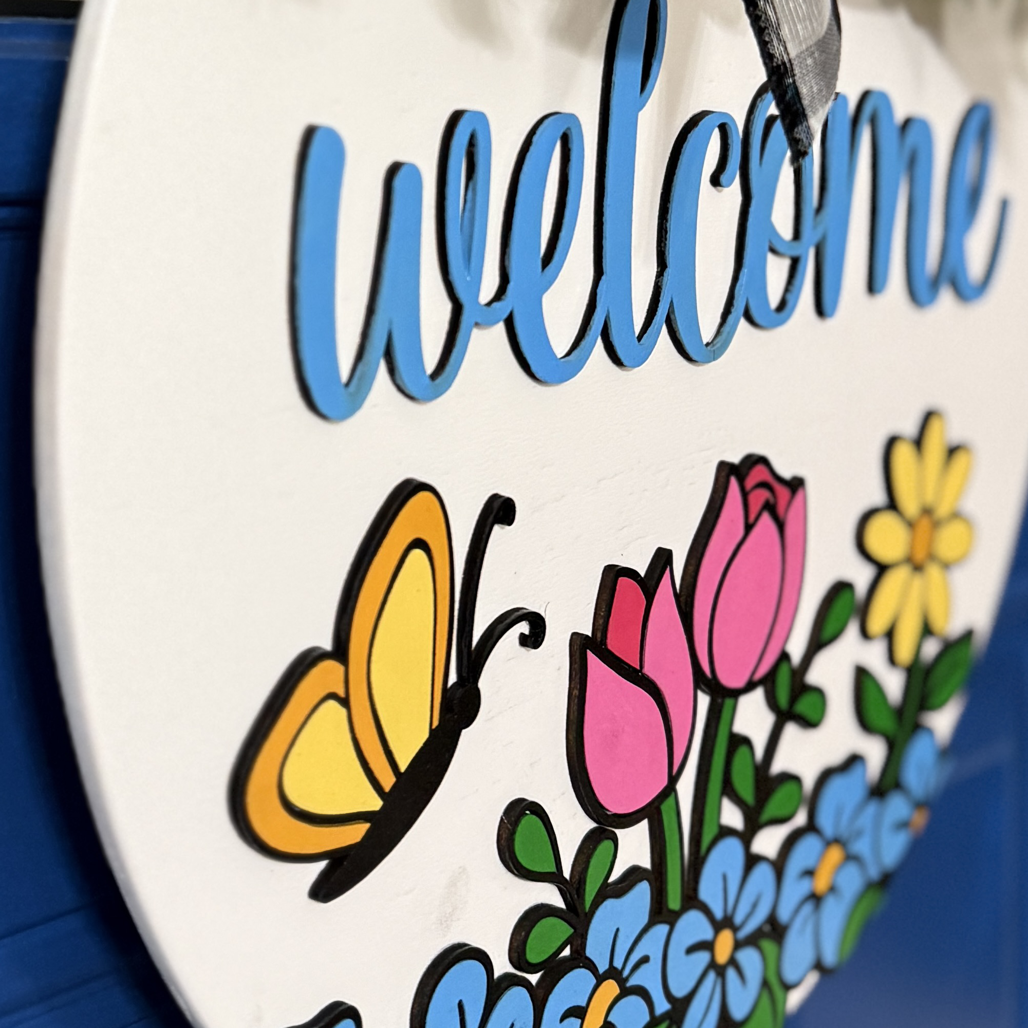 Close-up of yellow daisy, butterfly wing, and blue flowers on white round Welcome door hanger displayed on blue door