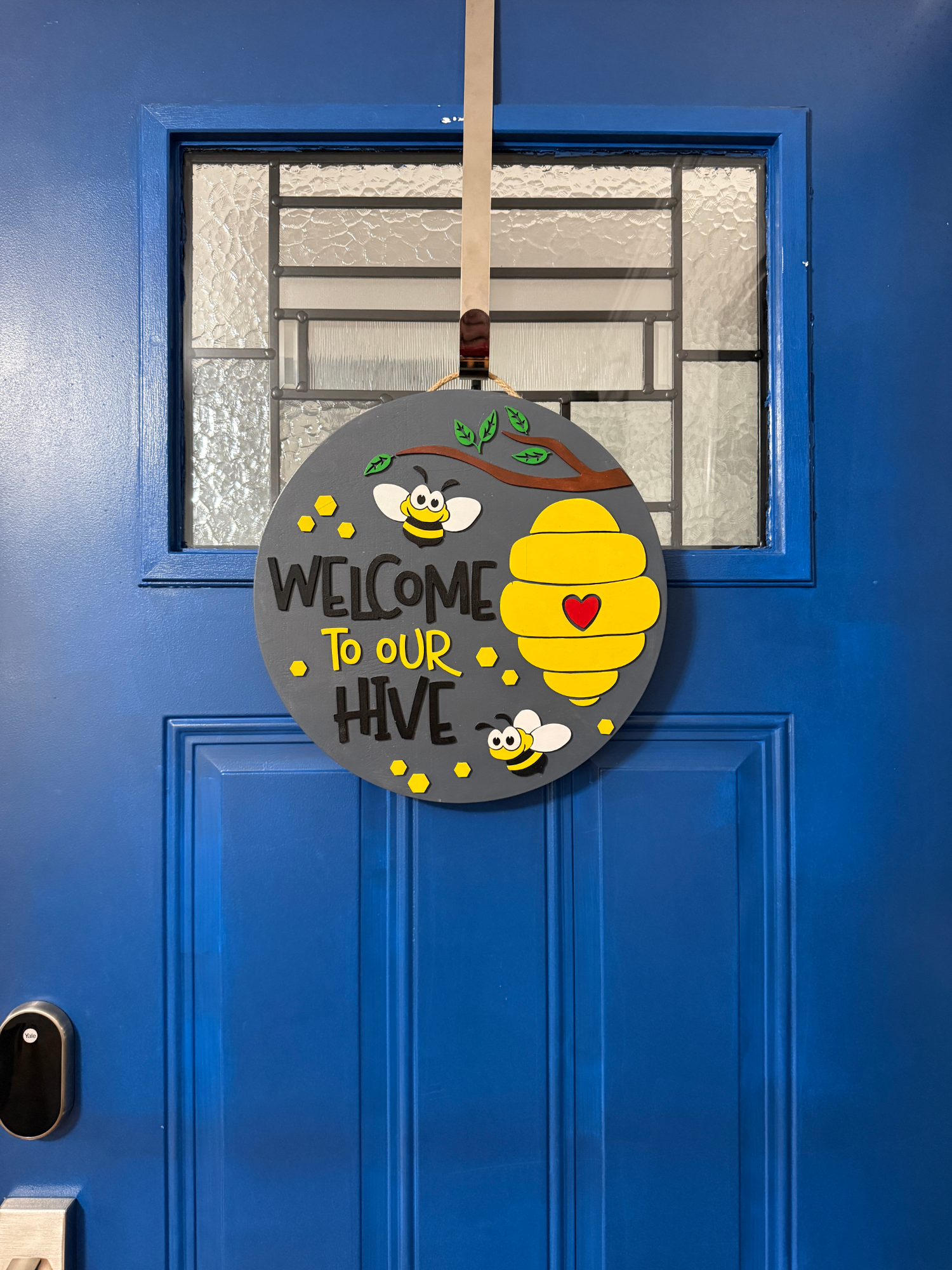 Front door display of gray bee hive-themed door hanger with beehive, bees, yellow accents, and “Welcome to Our Hive” text on a blue door