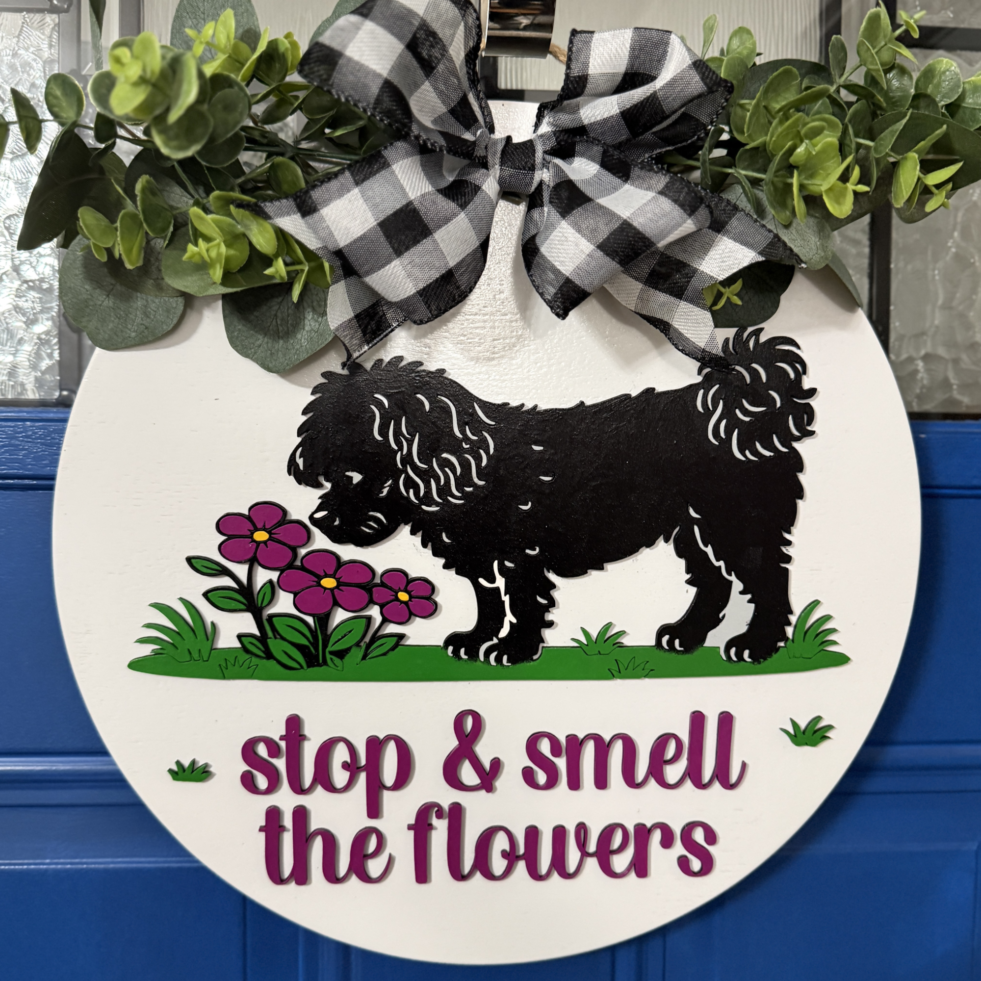 Front view of Stop and Smell the Flowers dog door hanger with black dog silhouette, pink flowers, magenta lettering, and buffalo check bow on blue door