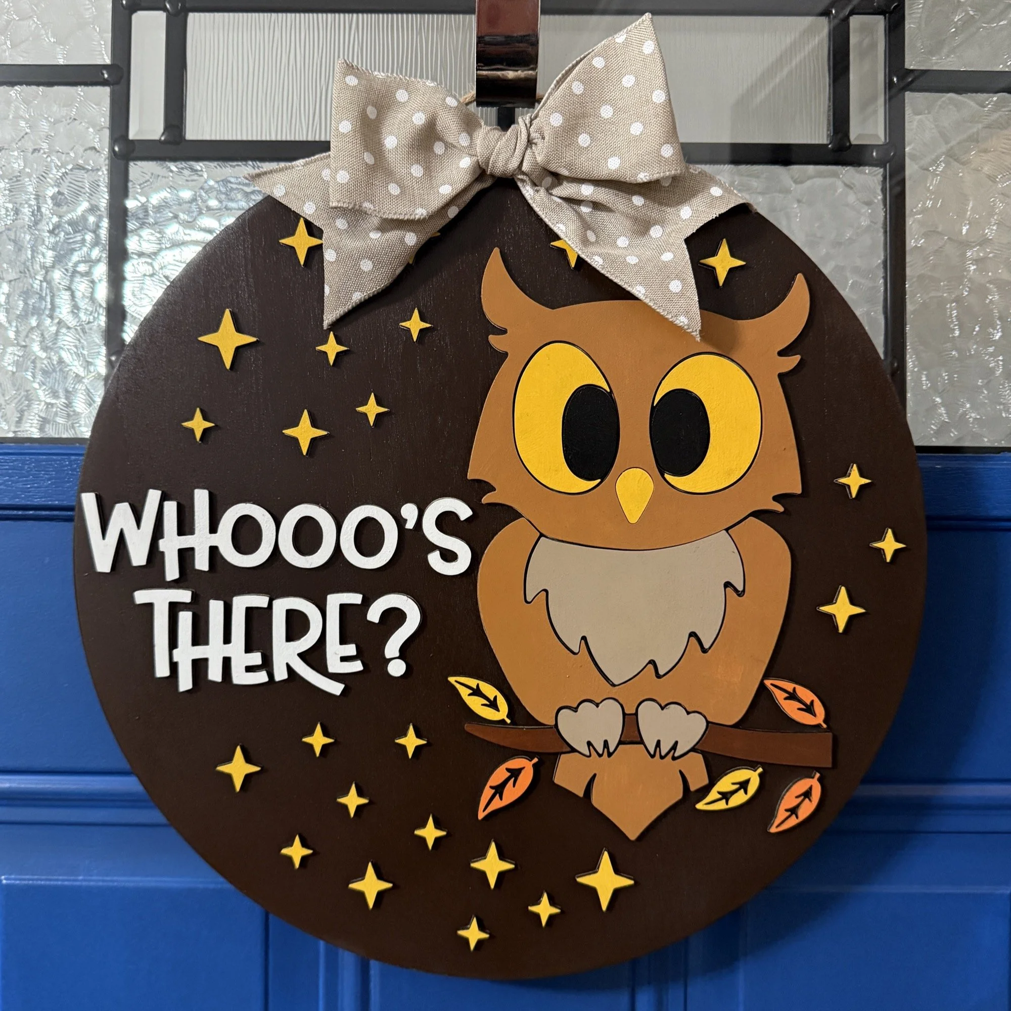 14-inch layered wood “Whooo’s There?” fall door hanger with owl, stars, and autumn leaves, topped with a polka dot bow, hanging on a blue front door.