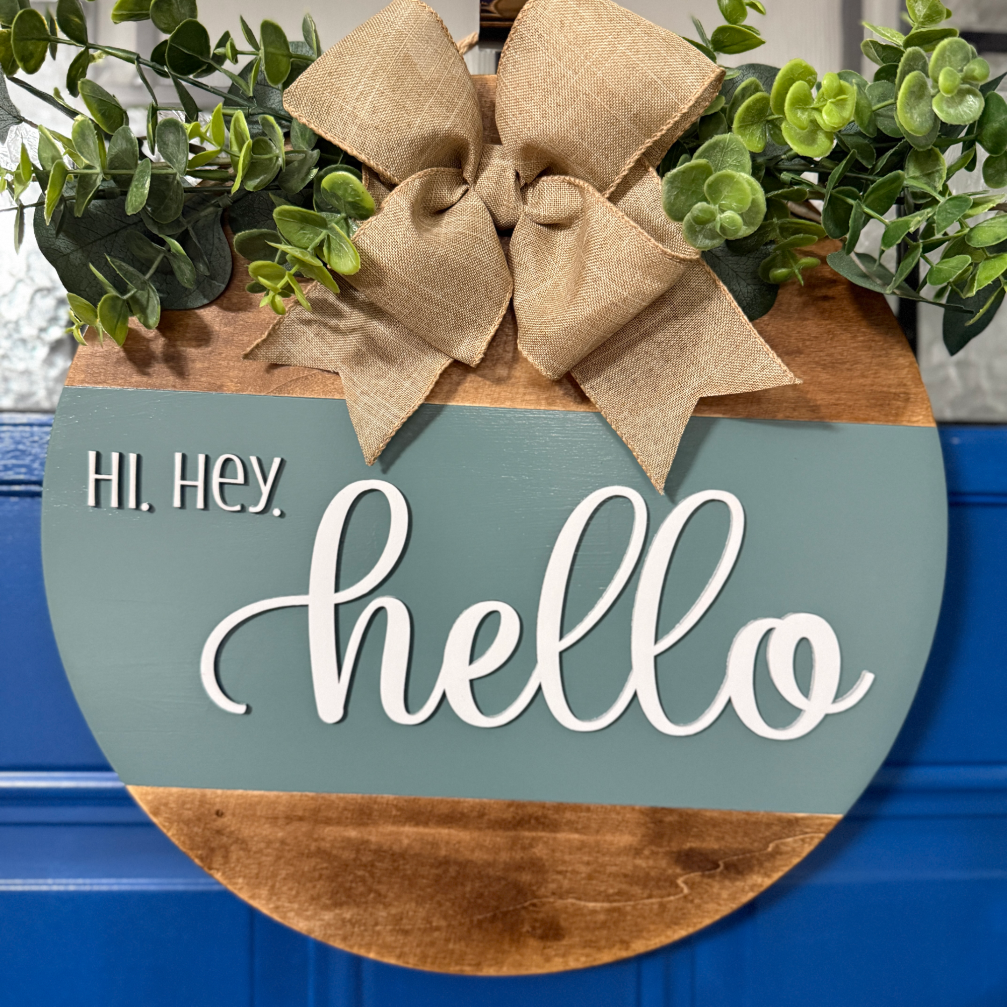 Sage green and stained wood hello door hanger with greenery accents and burlap bow hanging on blue front door