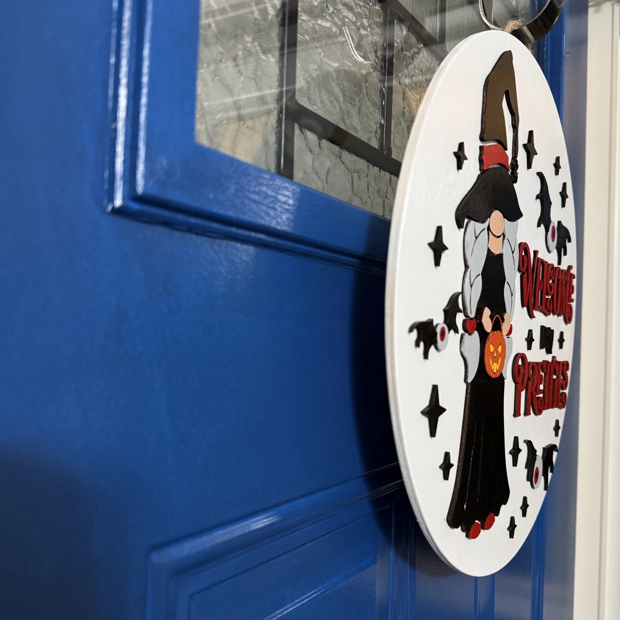 Side view of “Welcome My Pretties” Halloween door hanger with witch, bats, and stars on a white background, hanging on a blue front door.