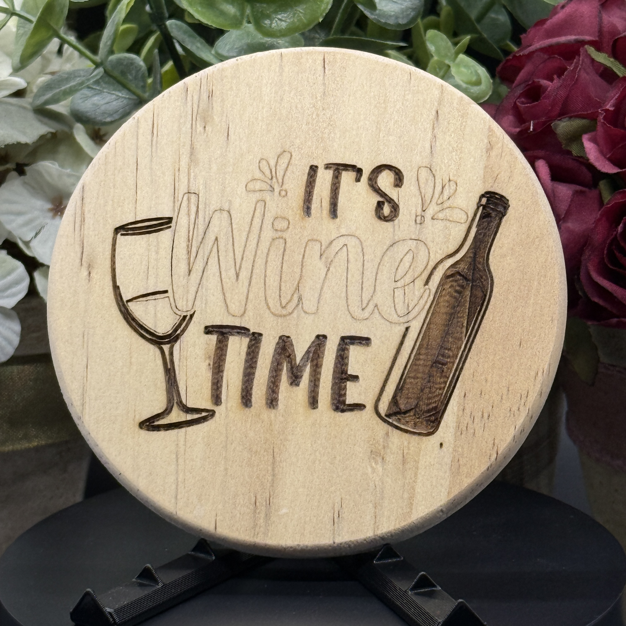 Round pine wood coaster with engraved wine bottle, glass, and the phrase ‘It’s Wine Time,’ displayed on a black stand with floral background