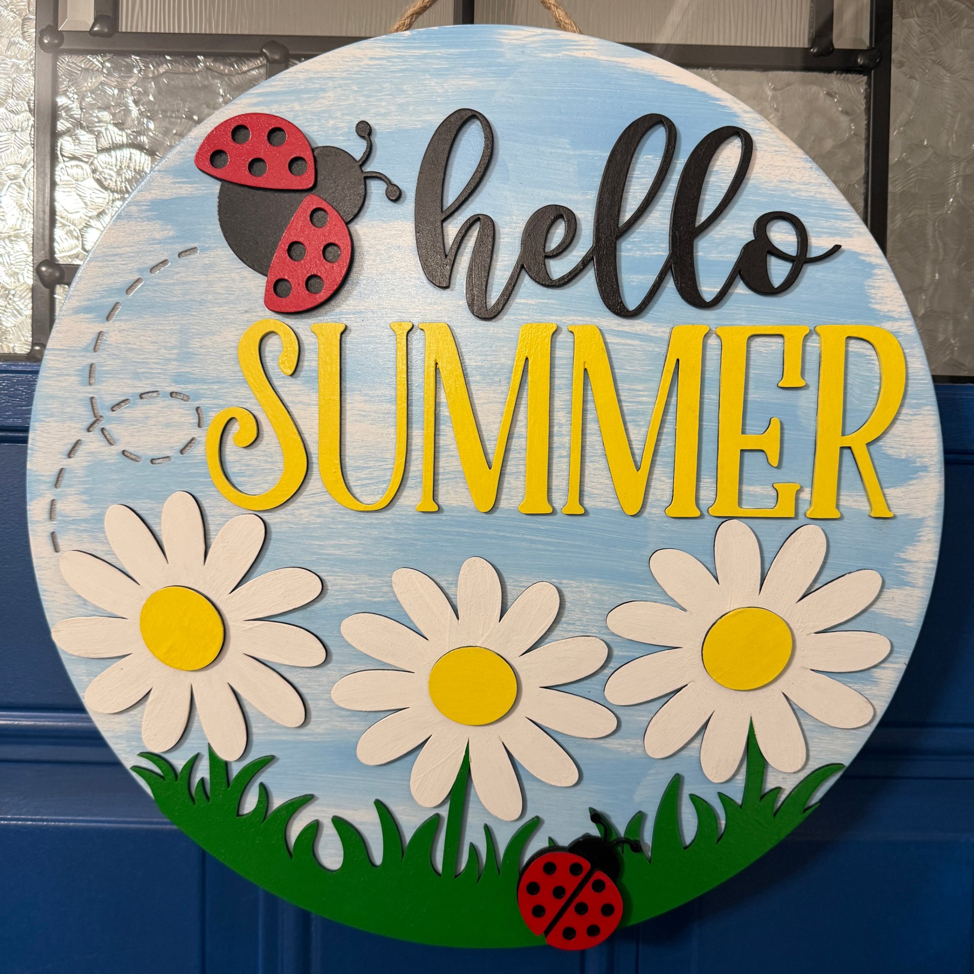 Front view of hello summer door hanger with yellow lettering, white daisies, and red ladybugs on a light blue wood background