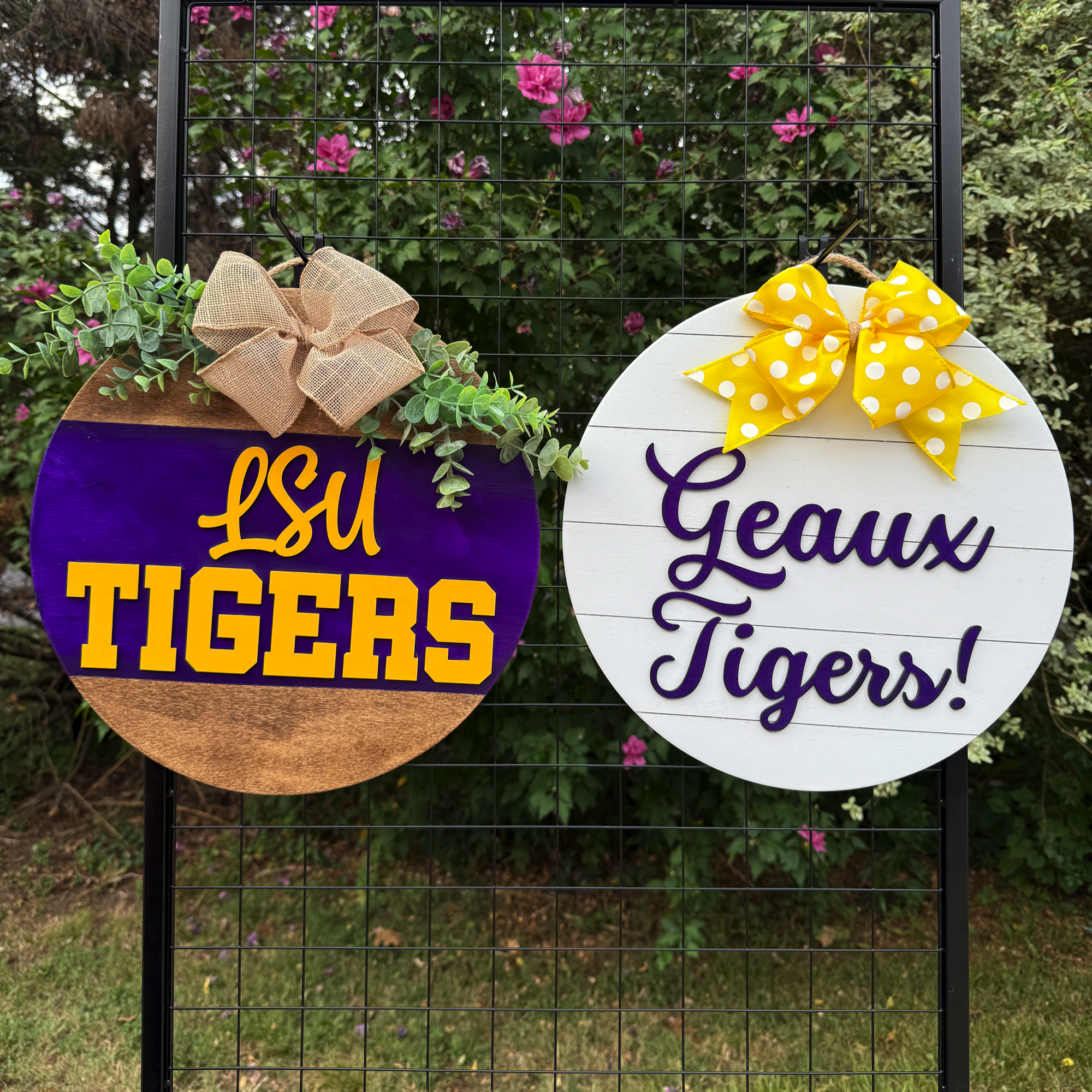 Two round LSU-themed door hangers displayed outdoors on a black grid with greenery in the background; one reads “LSU Tigers” and the other “Geaux Tigers!”