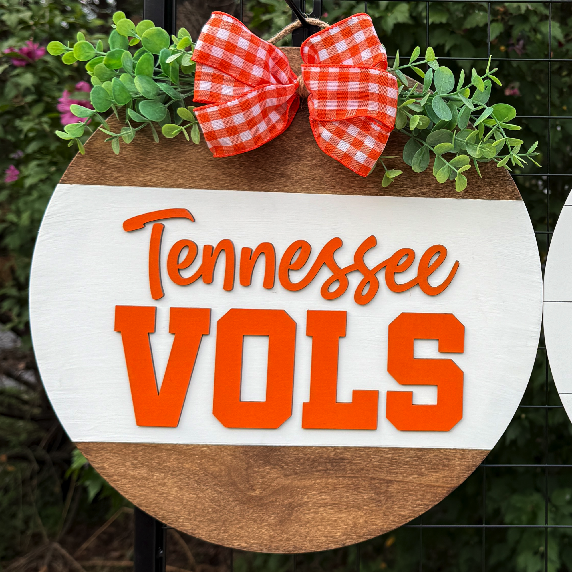 Round Tennessee Vols door hanger with bold orange lettering, greenery accents, and orange gingham bow on rustic wood background