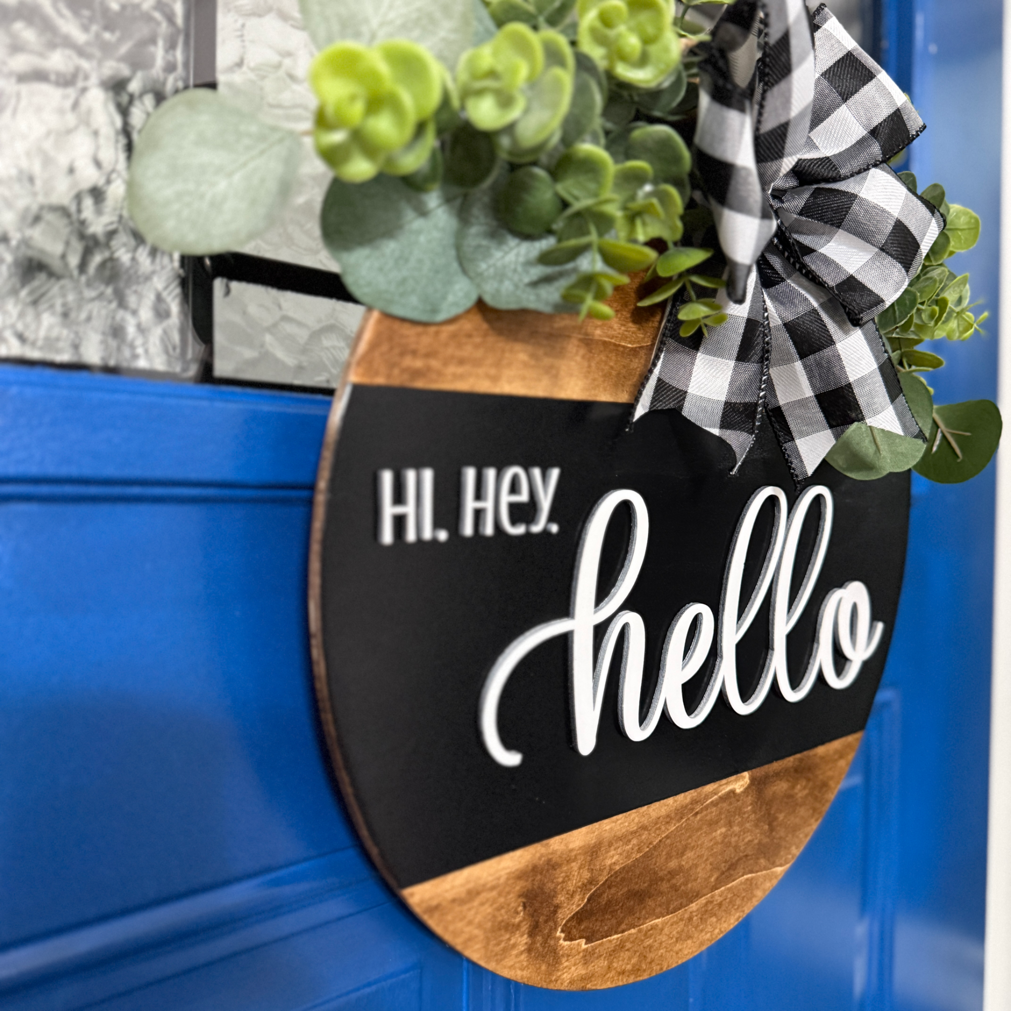 Angled view of black hello door hanger with buffalo plaid ribbon bow and eucalyptus greenery hanging on blue door