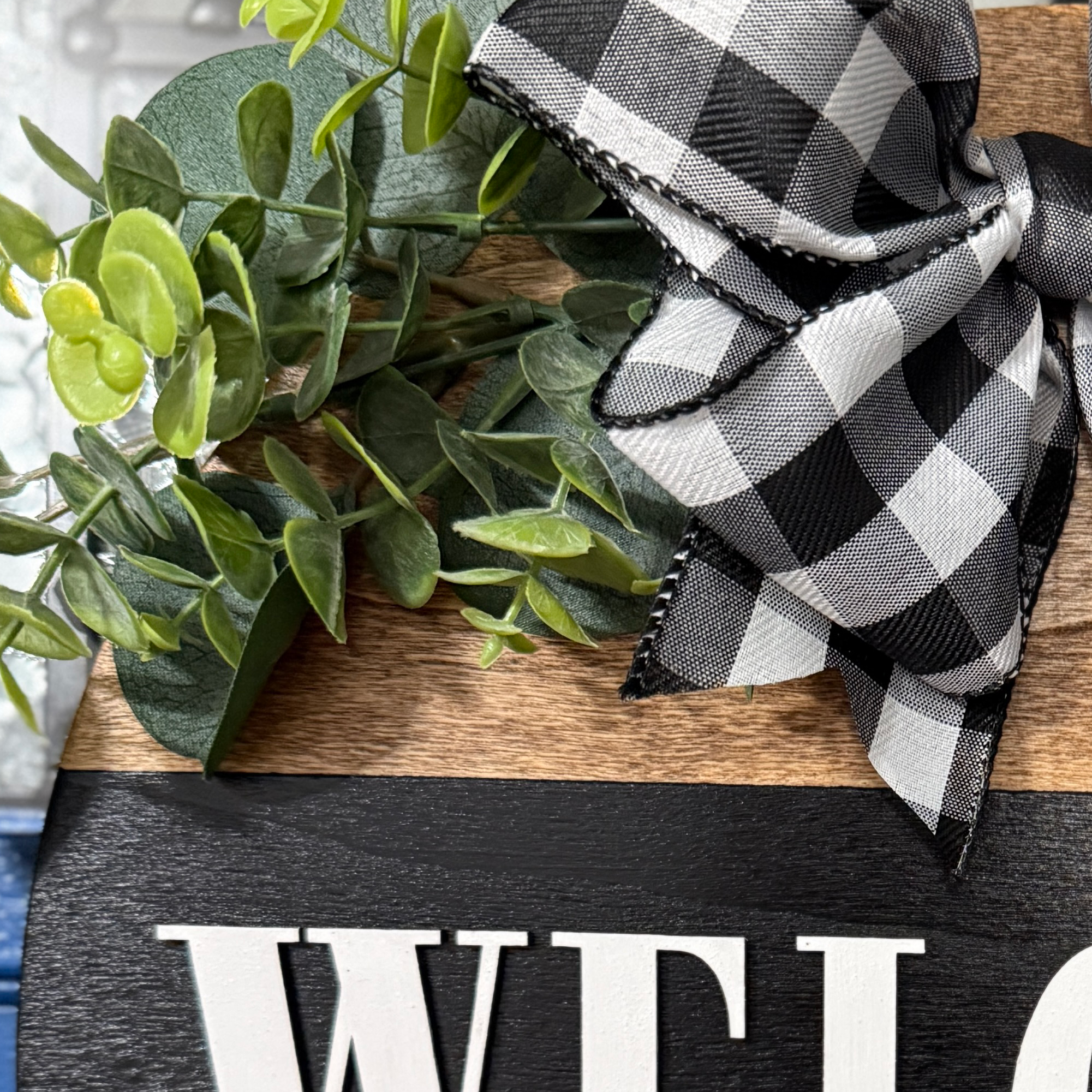 Close-up of faux greenery and black buffalo plaid bow attached to handcrafted welcome door sign