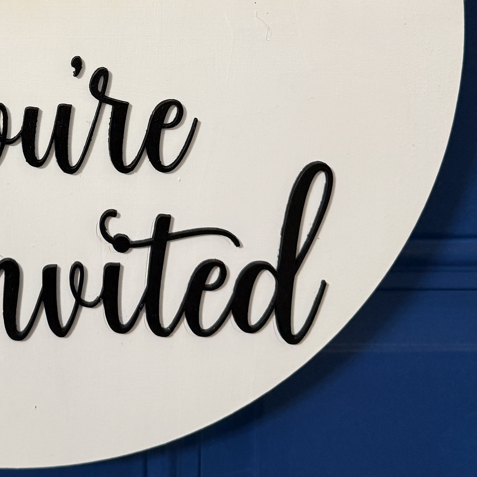 Close-up of raised black script lettering reading “You’re Uninvited” on round white wooden door hanger.