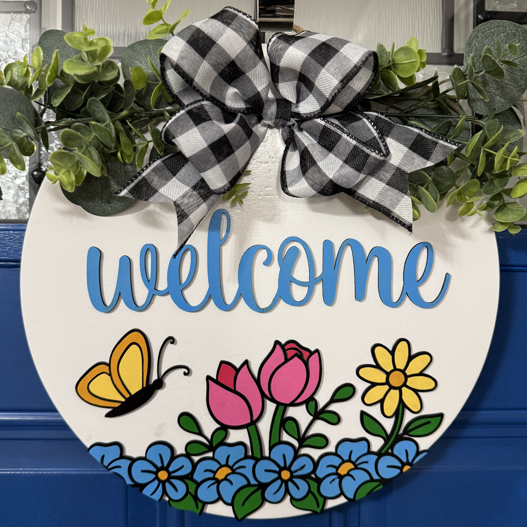 Front view of white round Welcome spring door hanger with blue raised lettering, colorful flowers, butterfly detail, and black and white buffalo check bow on blue door