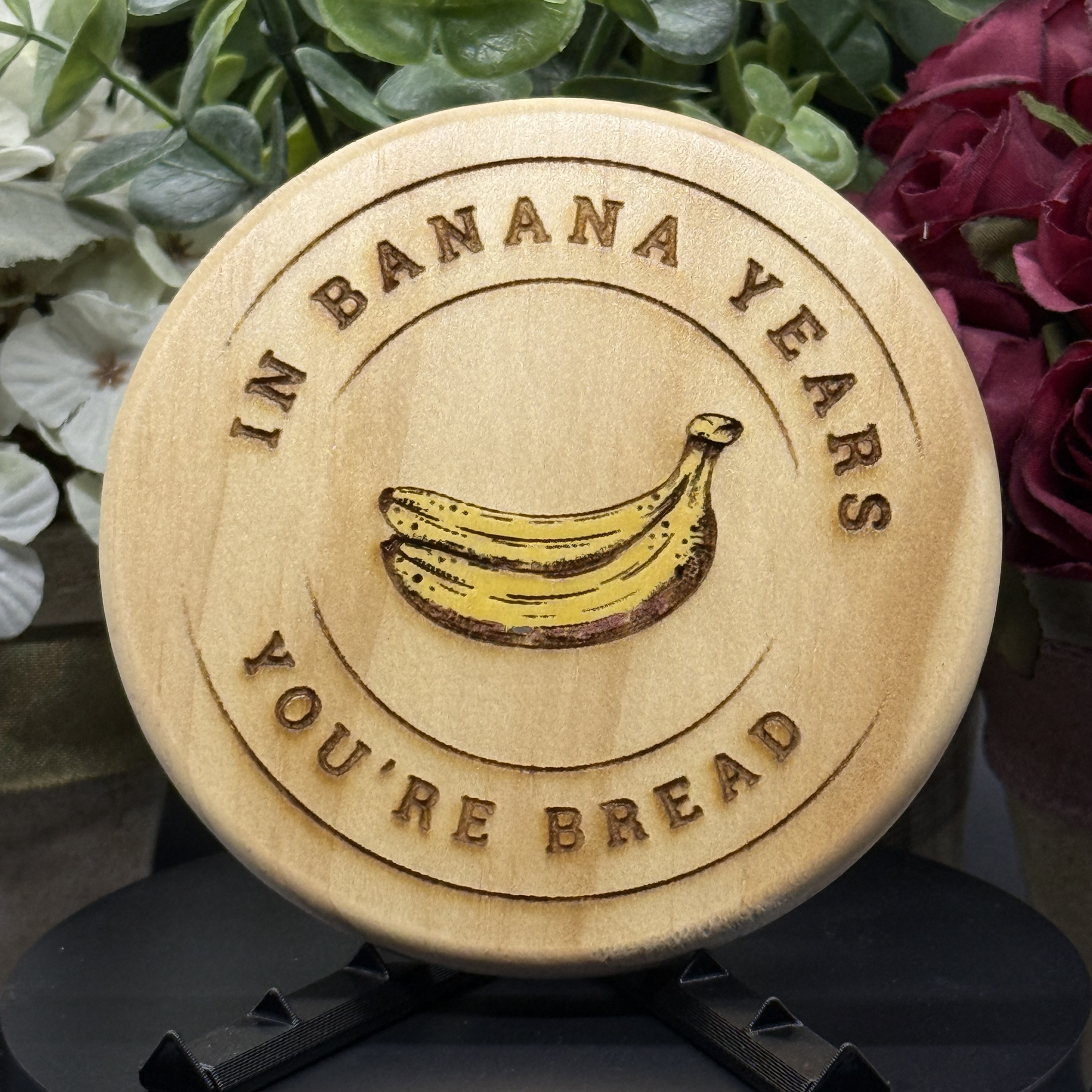 Round pine wood coaster with engraved and painted banana bunch and the phrase ‘In Banana Years, You’re Bread,’ displayed with floral background