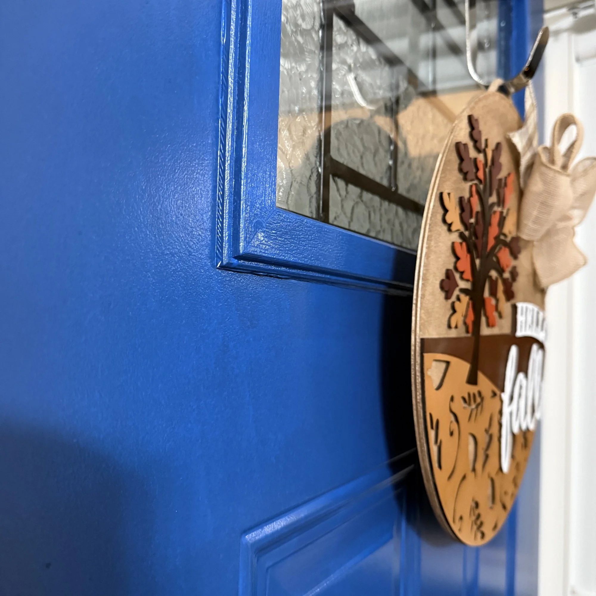 Side view of “Hello Fall” door hanger with autumn tree, orange and brown background, leaf cutouts, and burlap bow, hanging on a blue front door.