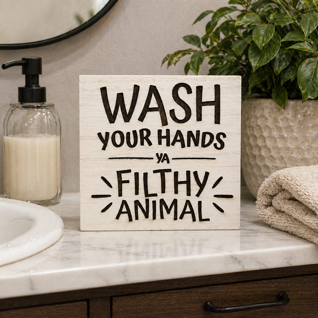 Bathroom shelf sitter sign reading “Wash your hands ya filthy animal” displayed on bathroom counter with soap dispenser, plant, and folded towels