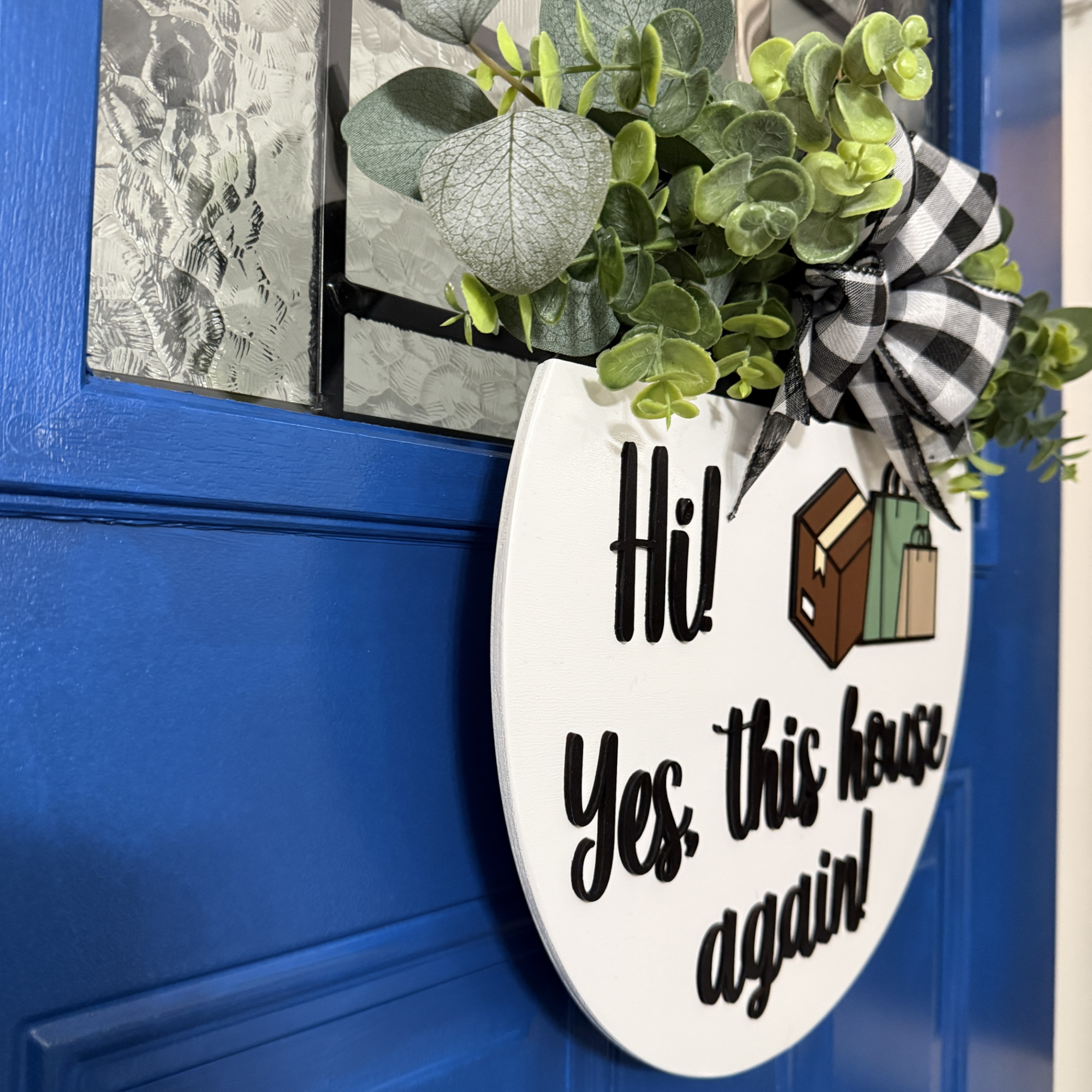 Angled side view of funny package delivery door hanger with raised black script lettering, box and shopping bag graphics, and buffalo plaid bow on a blue front door.