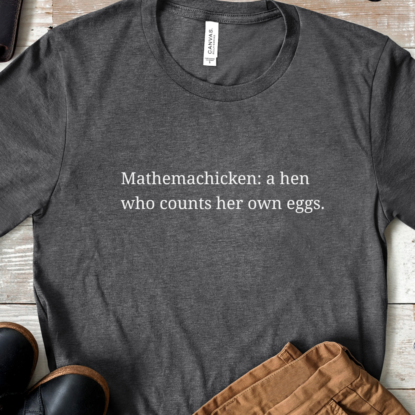 Charcoal gray t-shirt featuring the phrase “Mathemachicken: a hen who counts her own eggs” in soft white text, laid flat with casual outfit accessories