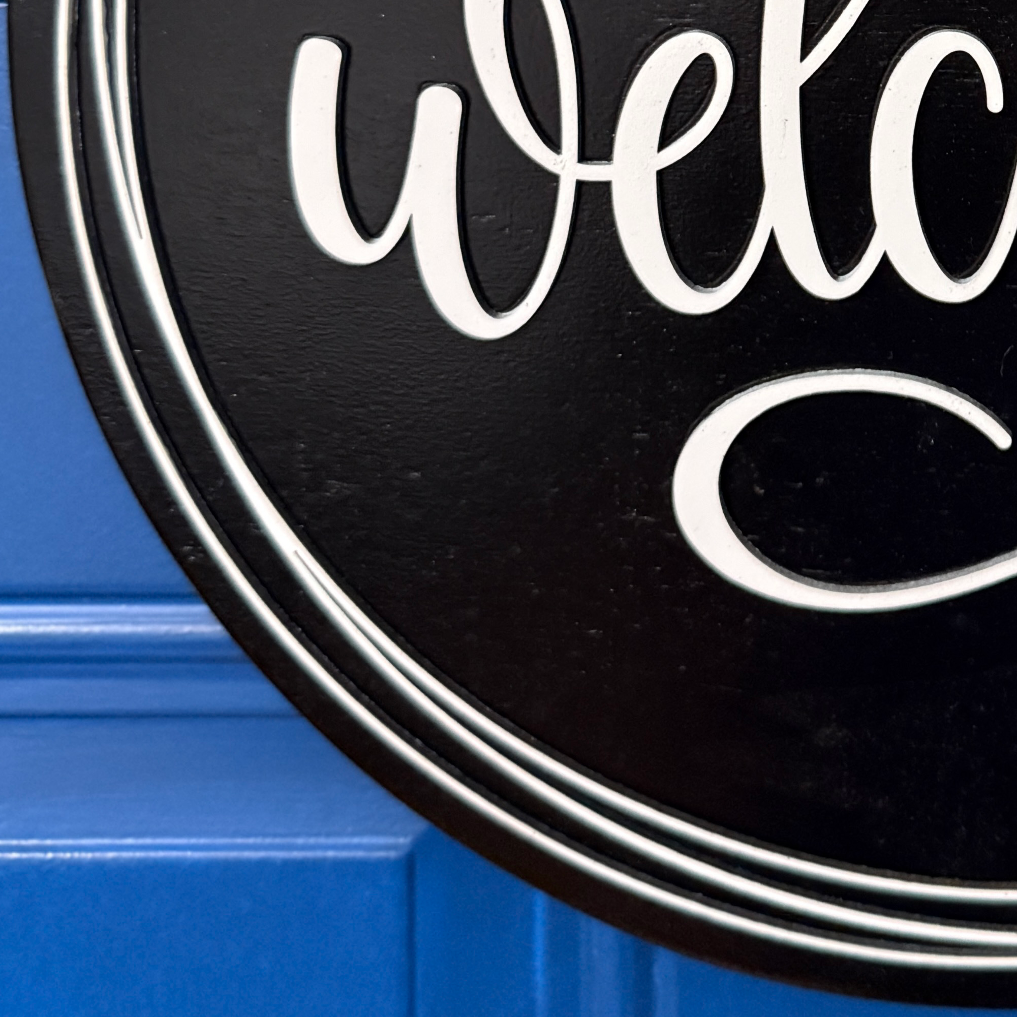 Close-up of white welcome script lettering on black painted wood door hanger with layered border detail