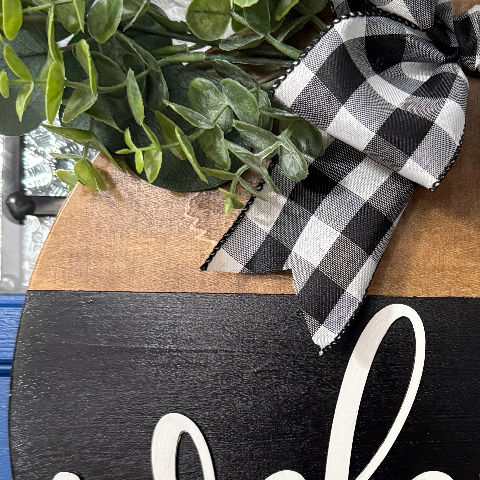 Close up of Welcome door hanger on stained base with black background, faux with greenery and coordinating bow