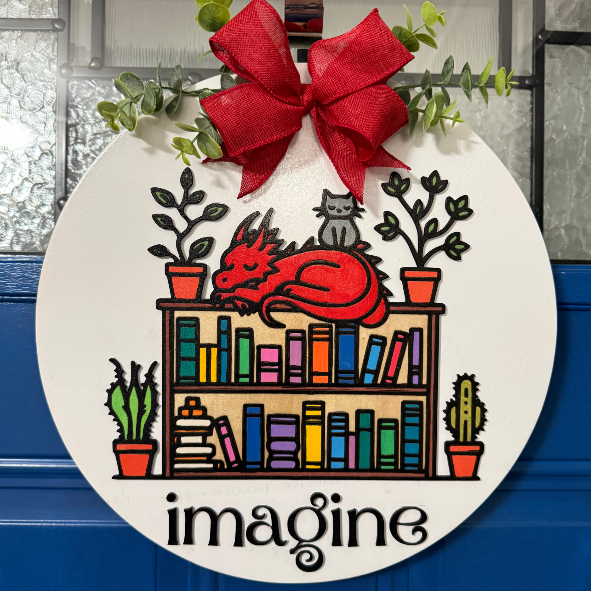 Front view of round wooden door hanger featuring red dragon resting on colorful bookshelf with “imagine” text and red bow