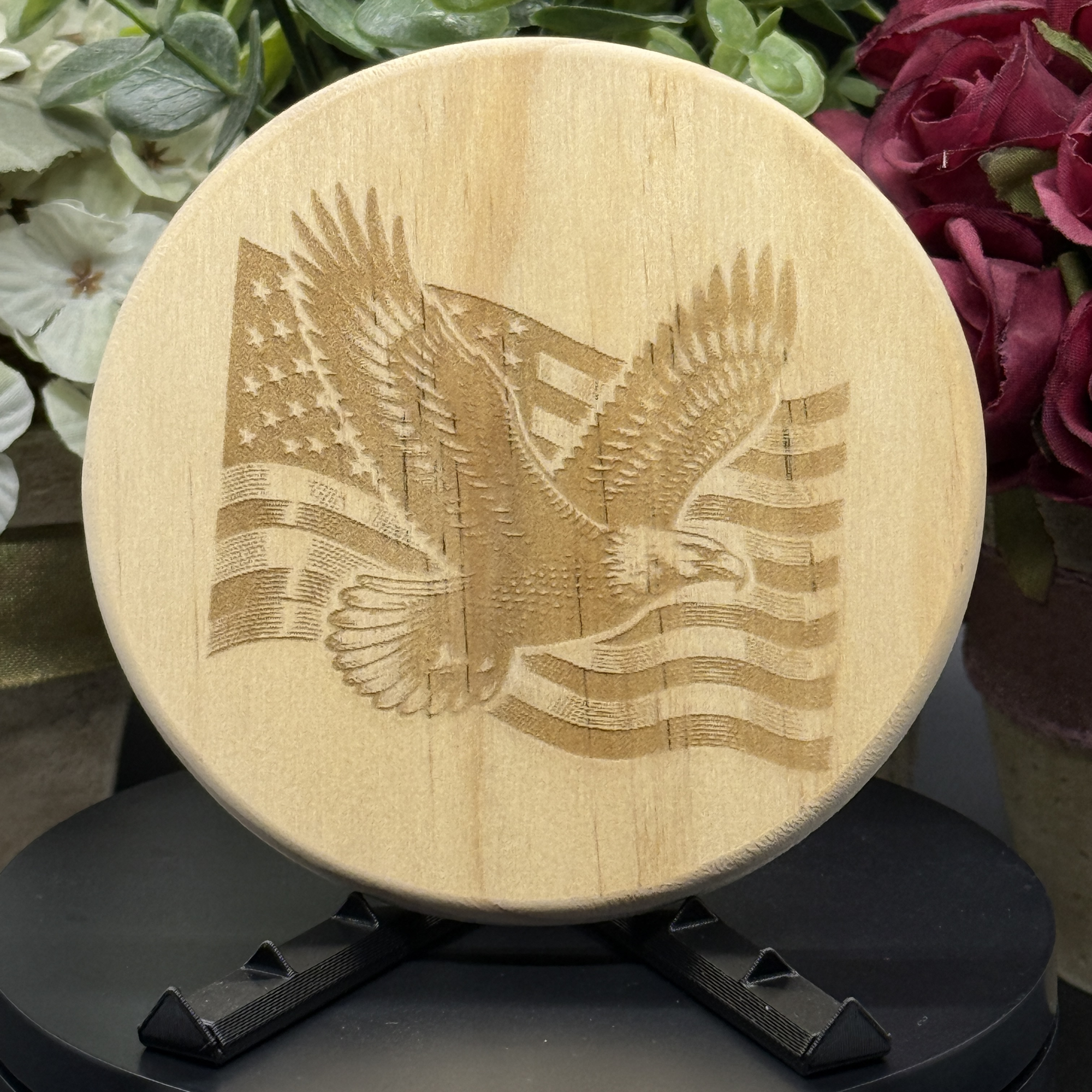 Round pine wood coaster with laser-engraved American flag and flying eagle design, displayed on a black stand with floral background