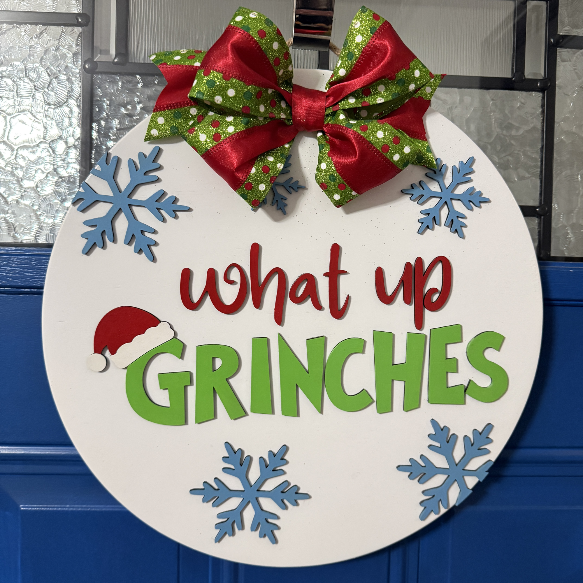 Front view of 14-inch wood door hanger featuring layered red and green “What Up Grinches” text with Santa hat accent, blue snowflakes, and red-green bow