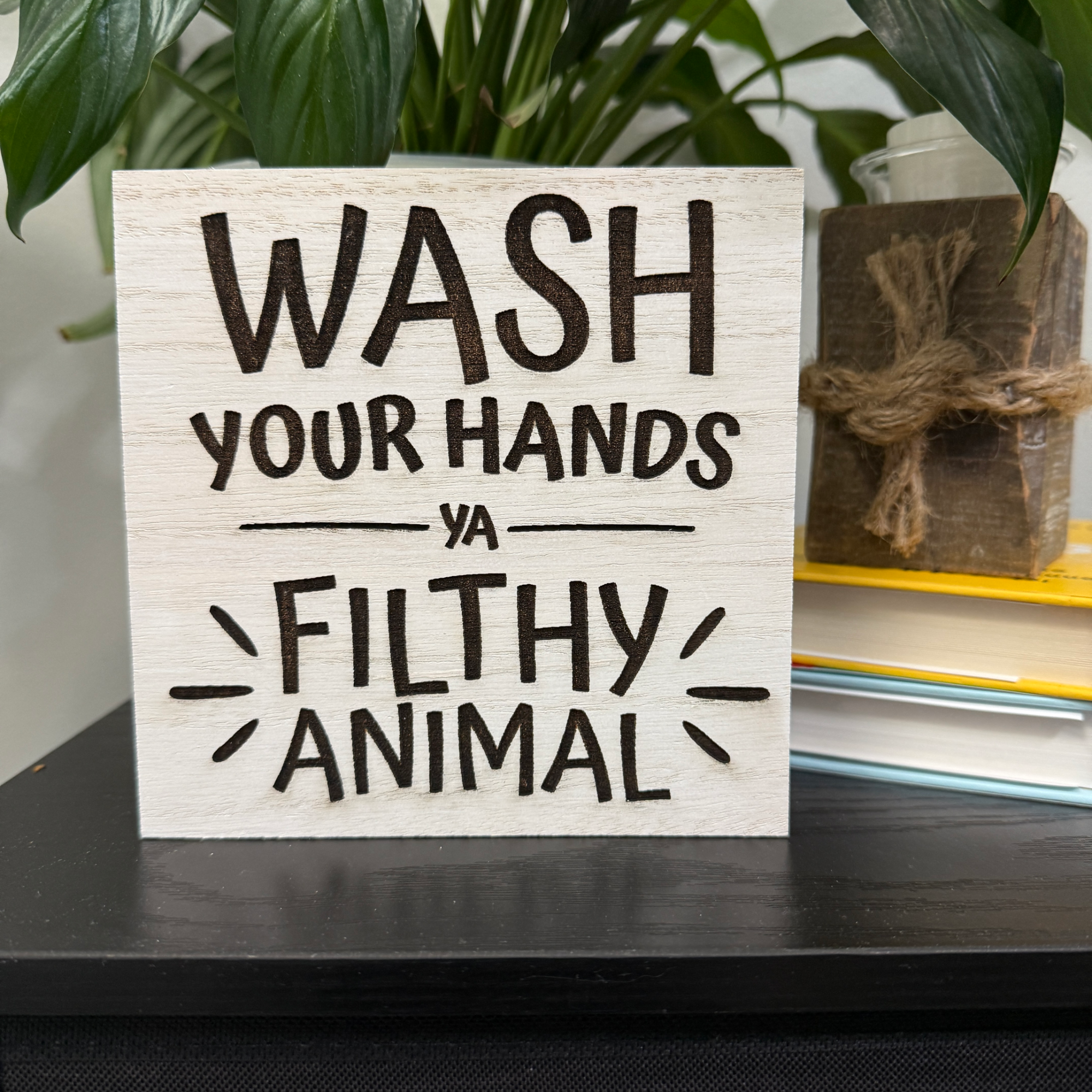 Close-up of humorous bathroom sign with bold engraved lettering on white-washed wood, styled beside greenery and stacked books