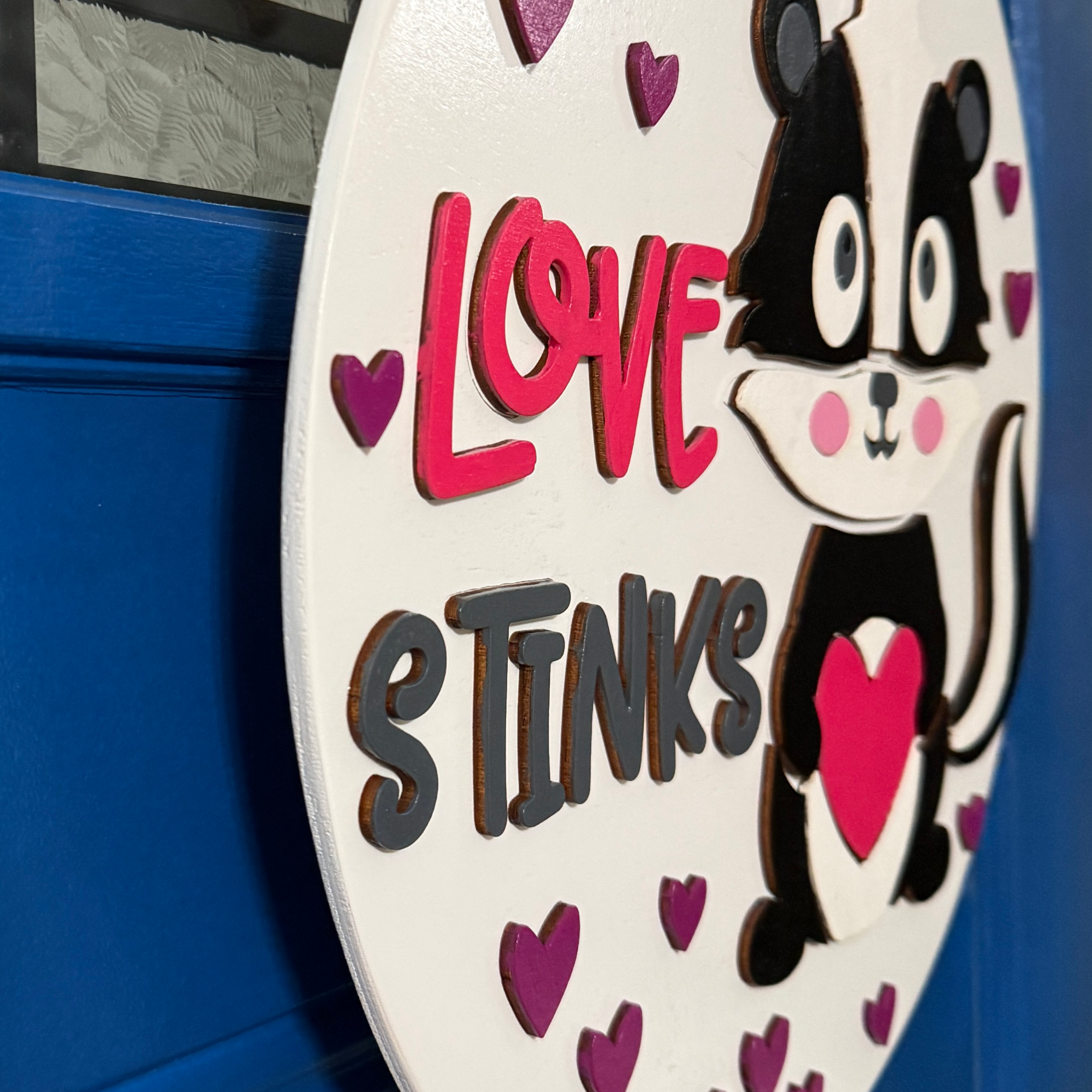 Angled close-up of Love Stinks Valentine door hanger showing raised lettering, hearts, and layered skunk design