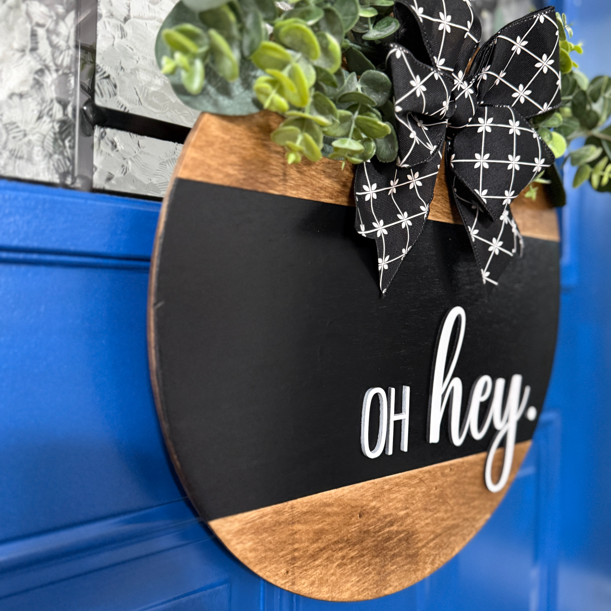 Angled view of black oh hey door hanger with eucalyptus greenery and black ribbon bow hanging on blue door