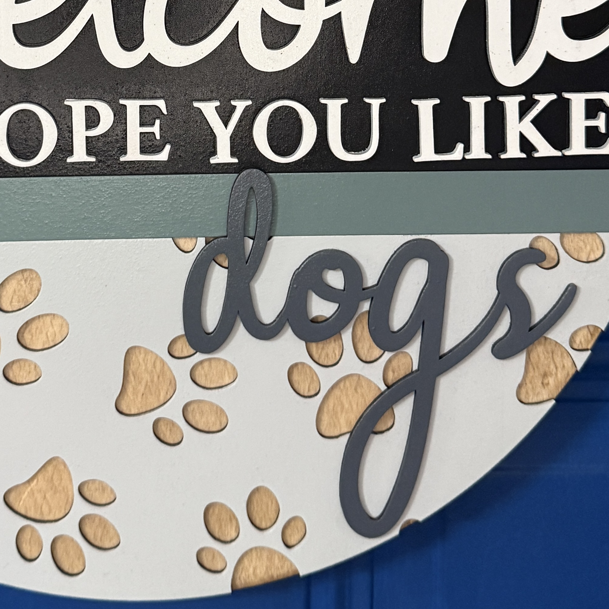 Close-up of “Welcome Hope You Like Dogs” door hanger with paw prints, heart accent, and neutral colors, hanging on a blue front door.