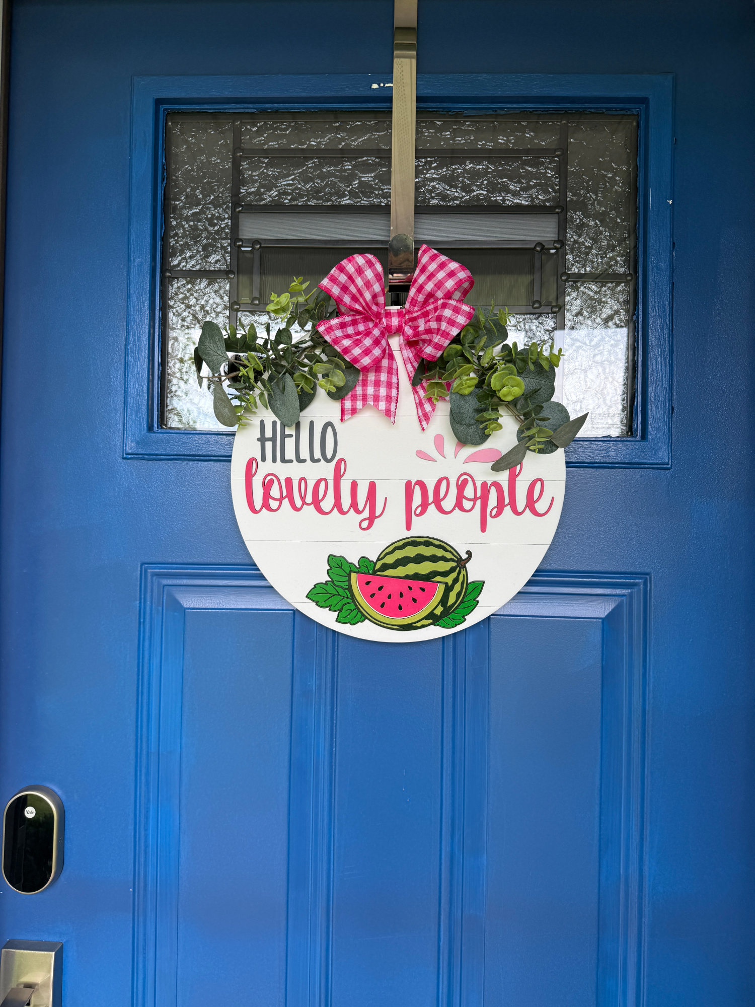 Watermelon-themed front door hanger displayed on a blue entry door with pink gingham bow, greenery, and Hello Lovely People greeting.