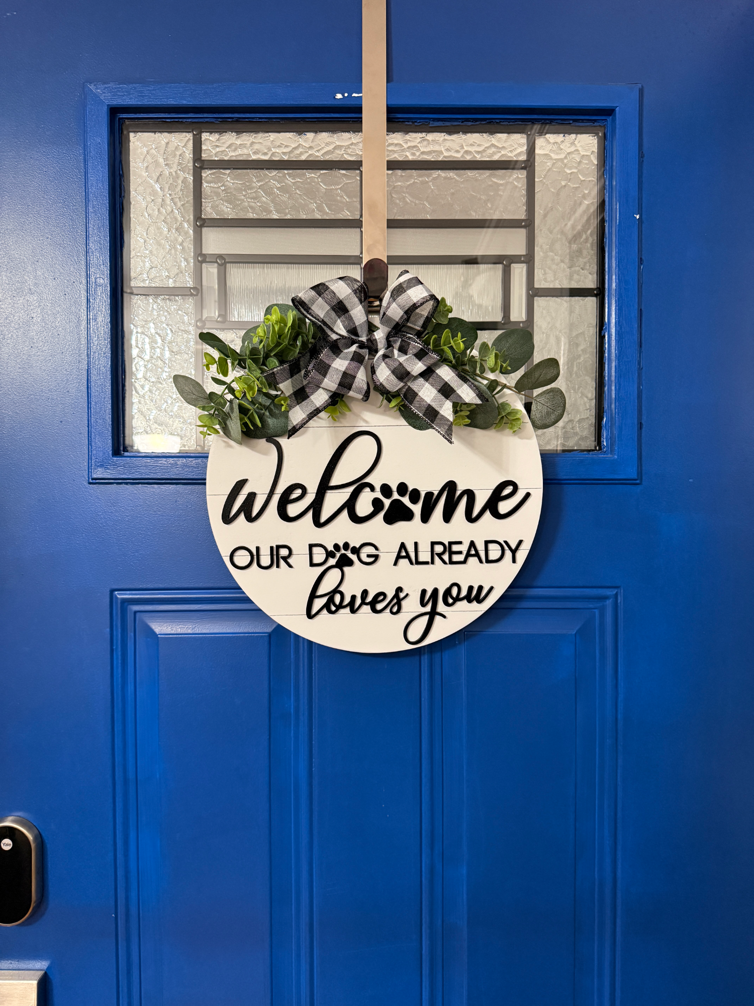 Front door display showing dog welcome sign centered on bright blue door with greenery accent