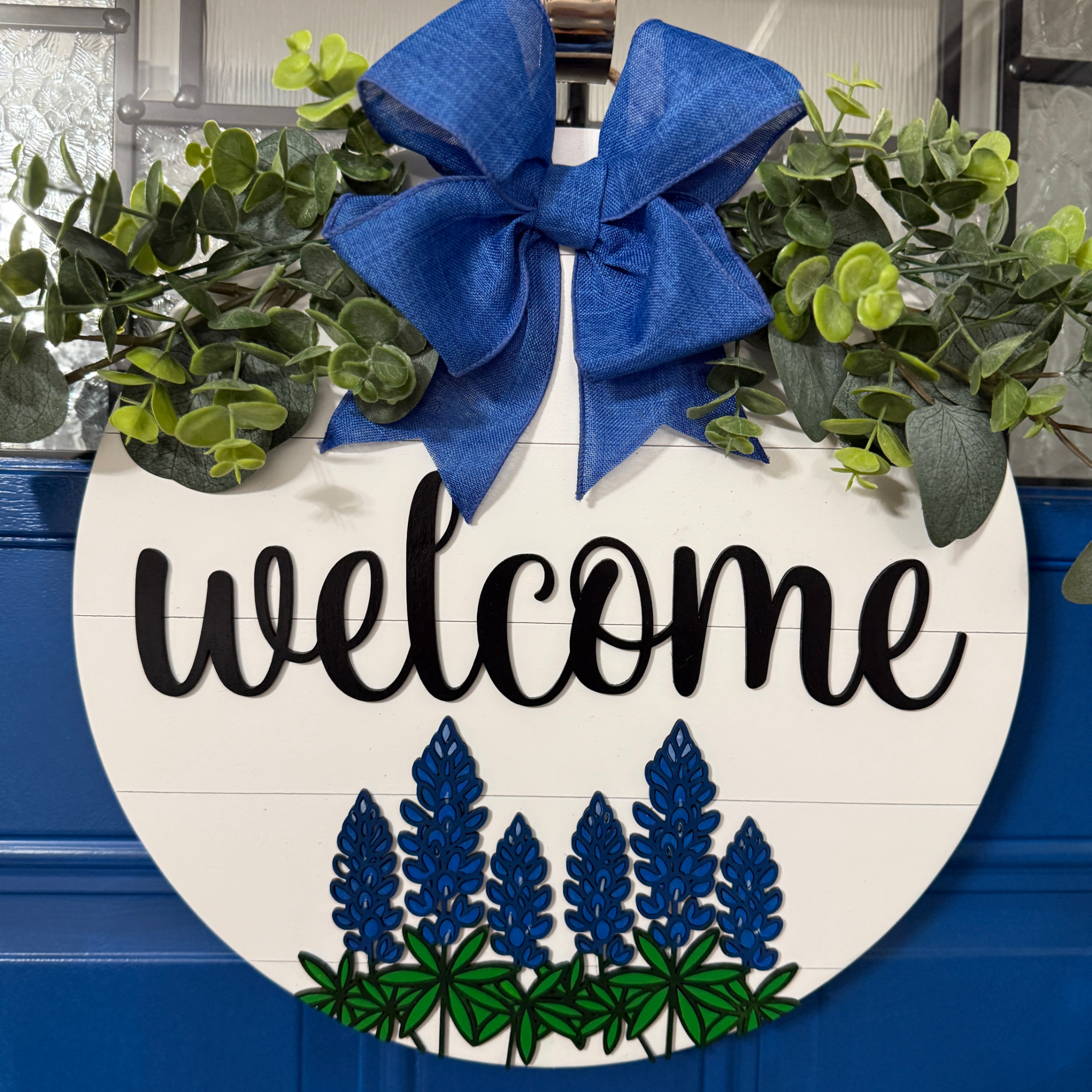 Bluebonnet welcome door hanger with blue bow and greenery on white shiplap background