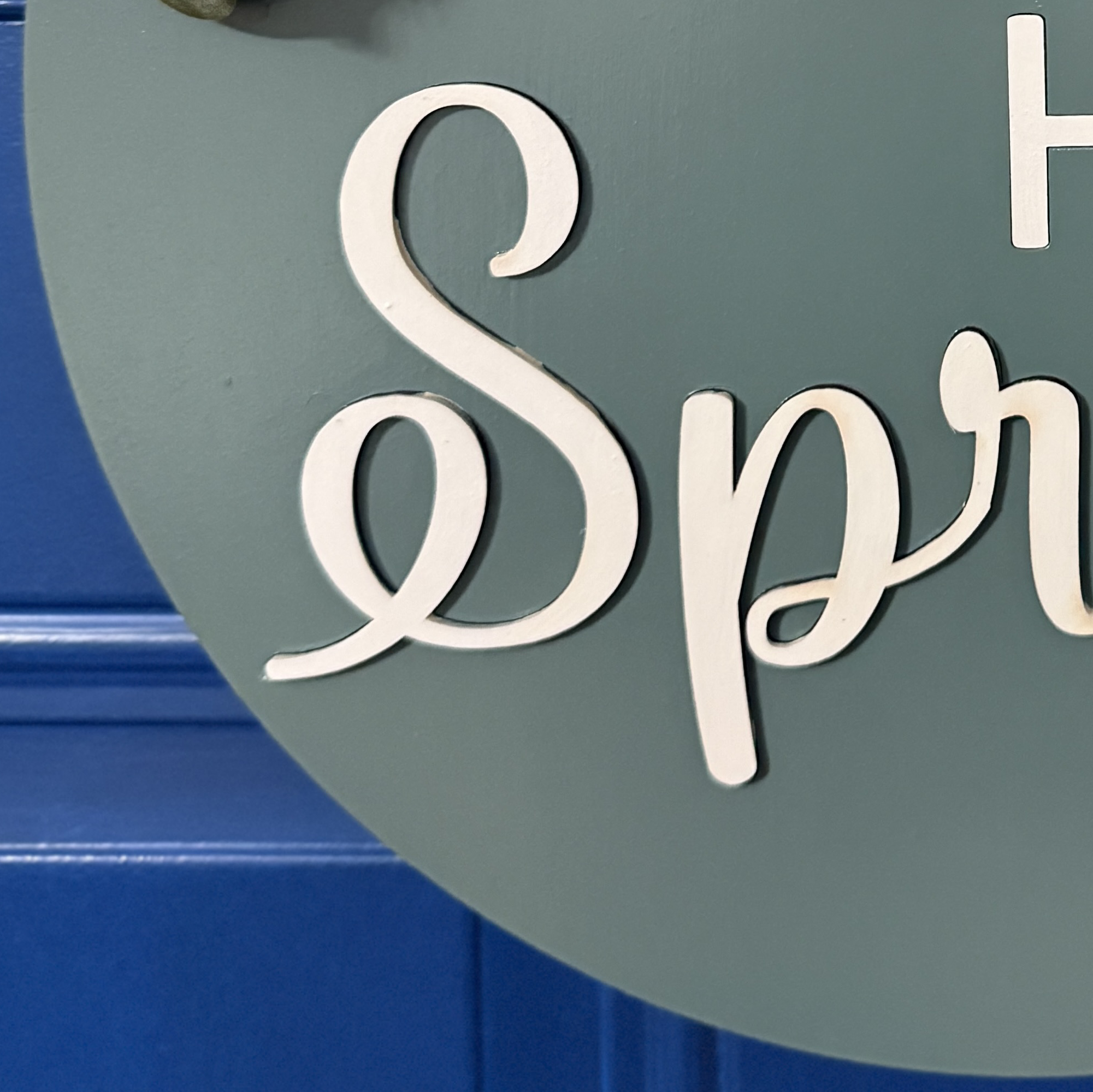 Close-up of white raised “Spring” script lettering on patina blue round wood door hanger