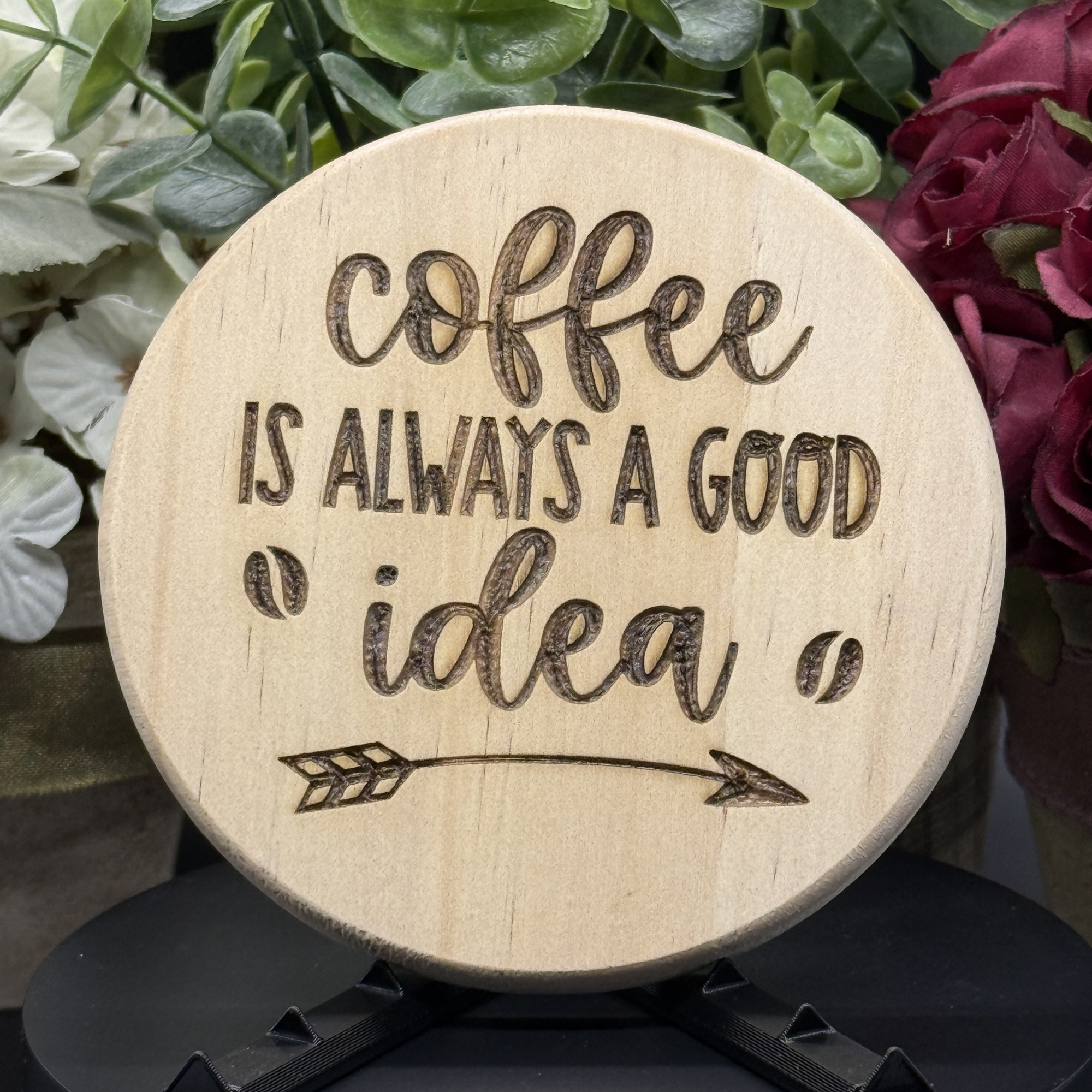 Round pine wood coaster with engraved phrase ‘Coffee Is Always a Good Idea’ and arrow detail, shown with floral background