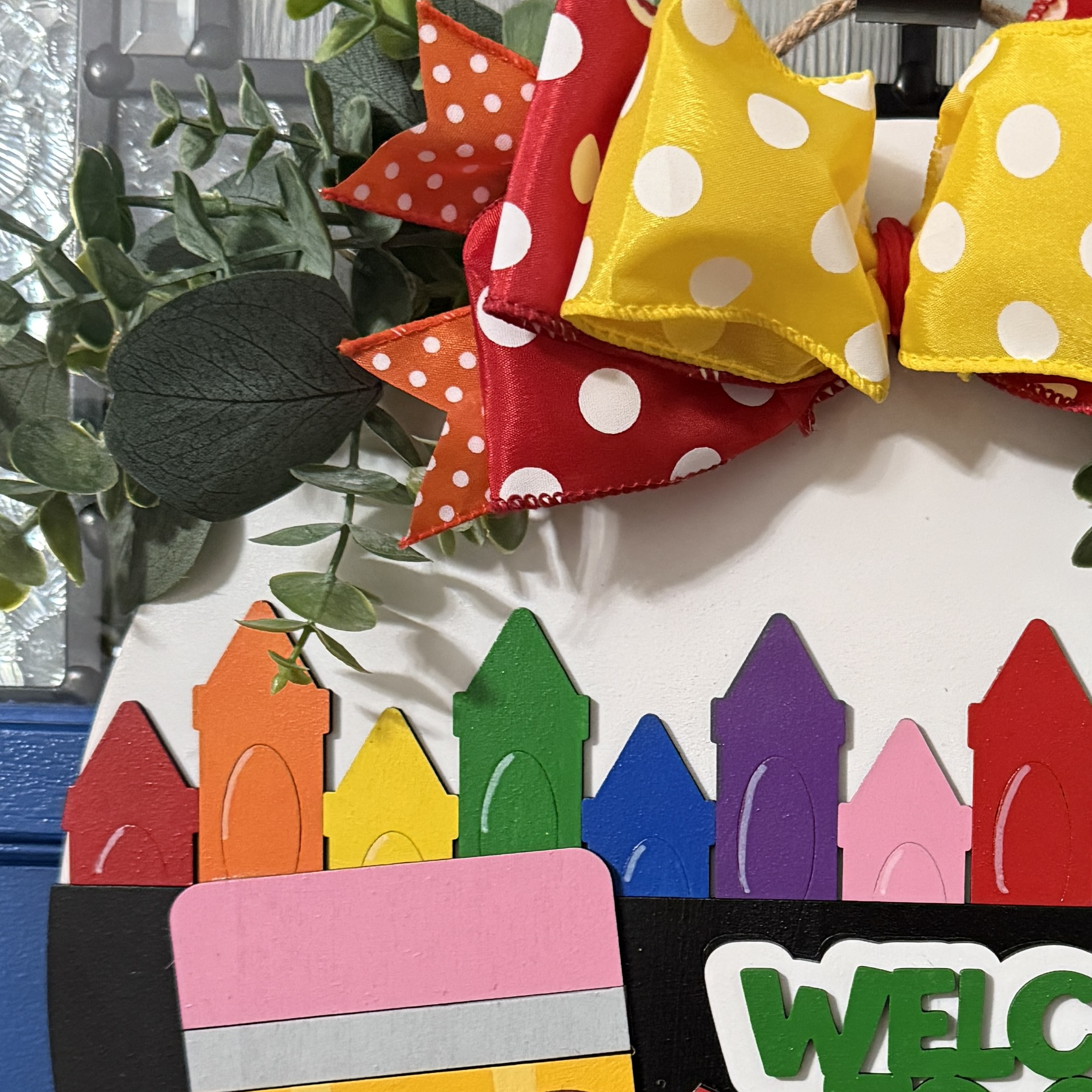 Side view of “Welcome to Our Class” door hanger featuring smiling pencil, rainbow crayon row, and red/yellow polka dot bow with greenery on a dark blue door.