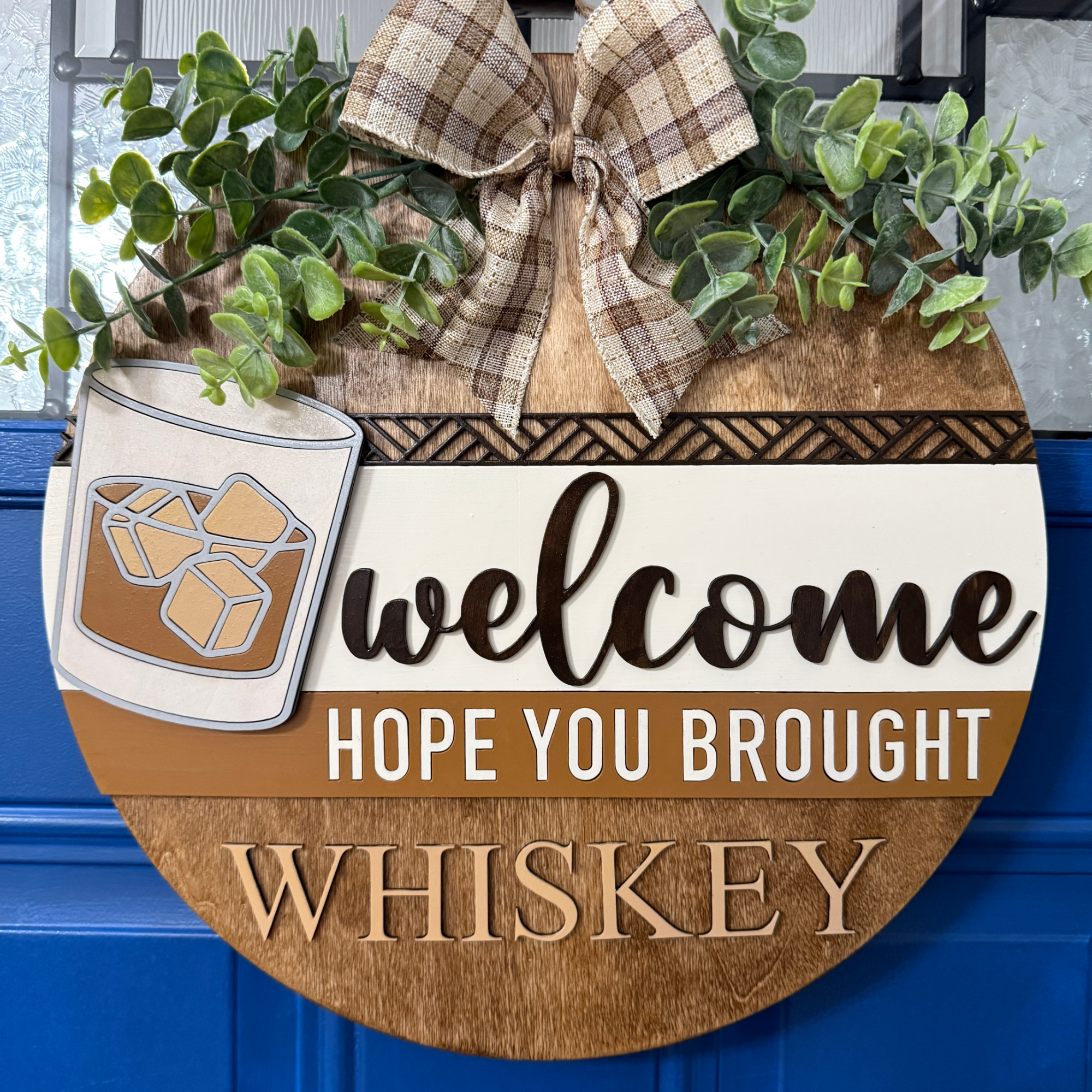 Front view of round wooden whiskey welcome door hanger with glass of whiskey, greenery, and “Hope you brought whiskey” text