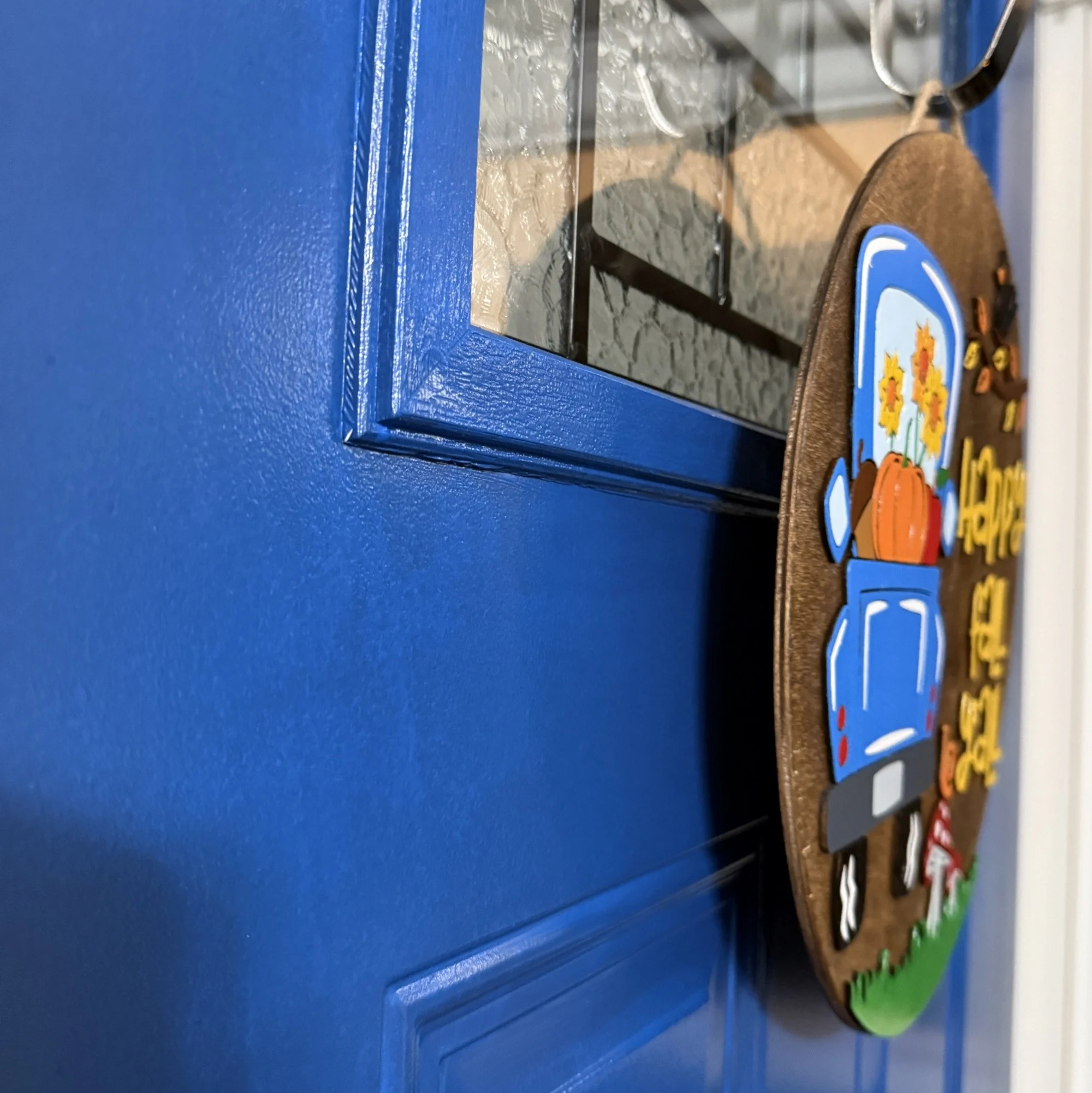Side view of “Happy Fall Y’all” door hanger with blue truck carrying pumpkins and sunflowers, autumn leaves, mushrooms, and a snail, hanging on a blue front door.