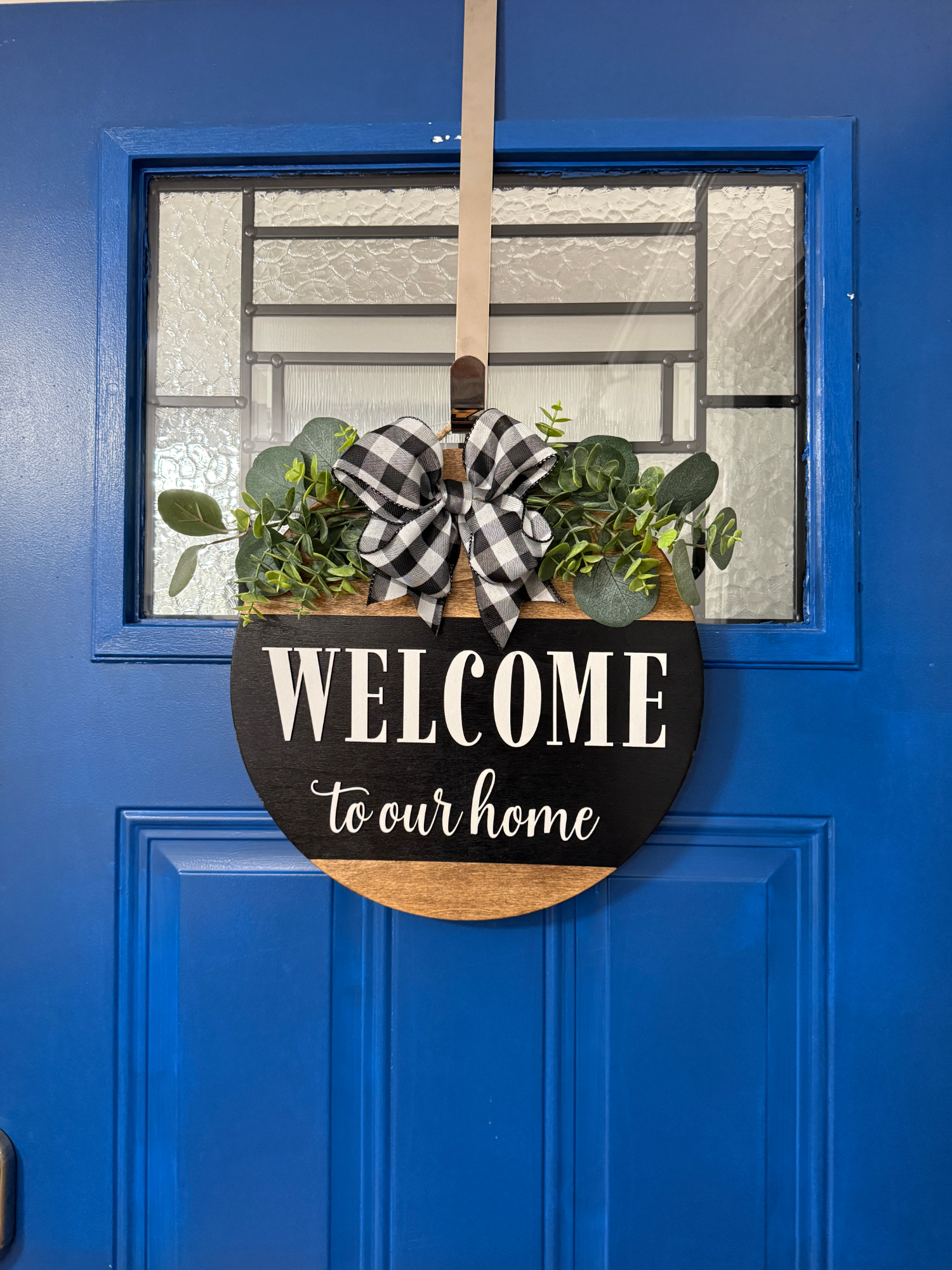 Full front door display featuring black welcome to our home door hanger with greenery and buffalo plaid bow