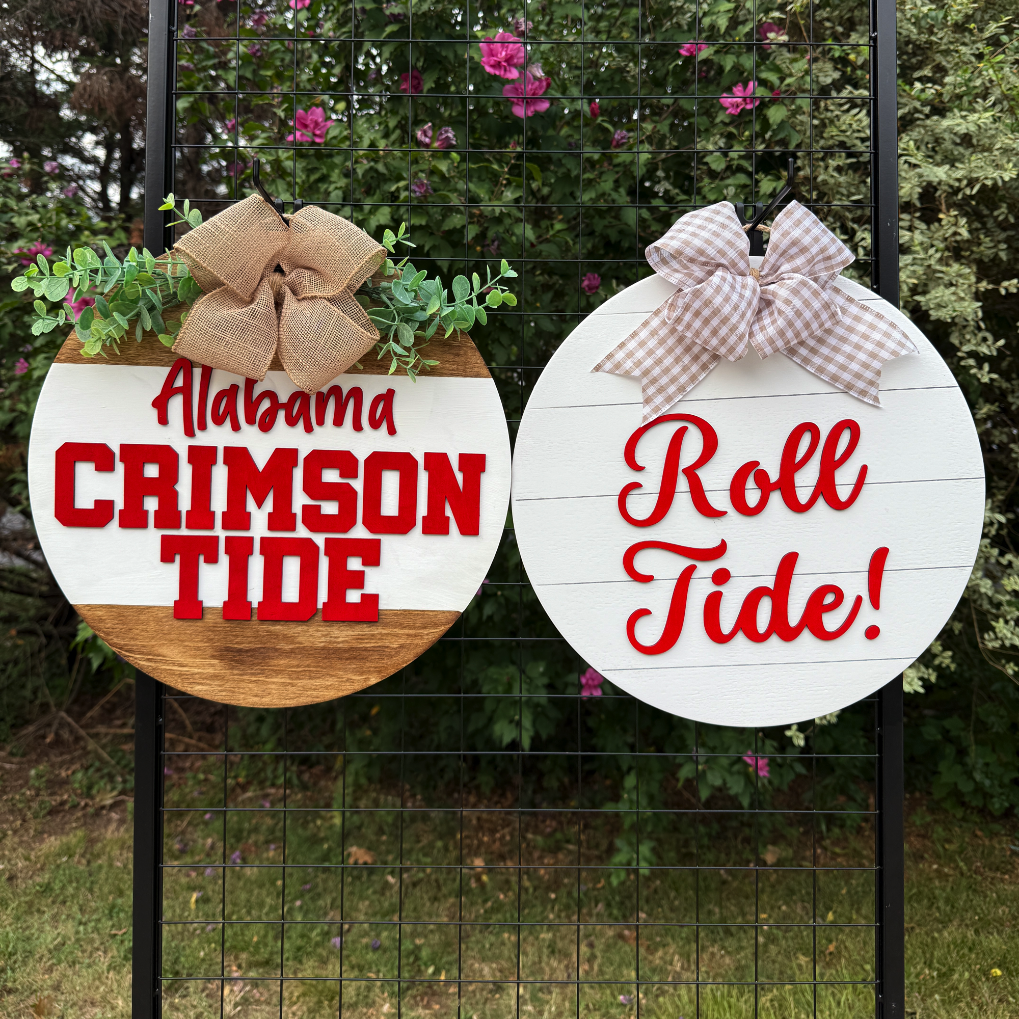 Two round Alabama-themed door hangers displayed outdoors on a black grid with greenery in the background; one reads “Alabama Crimson Tide” and the other “Roll Tide!”