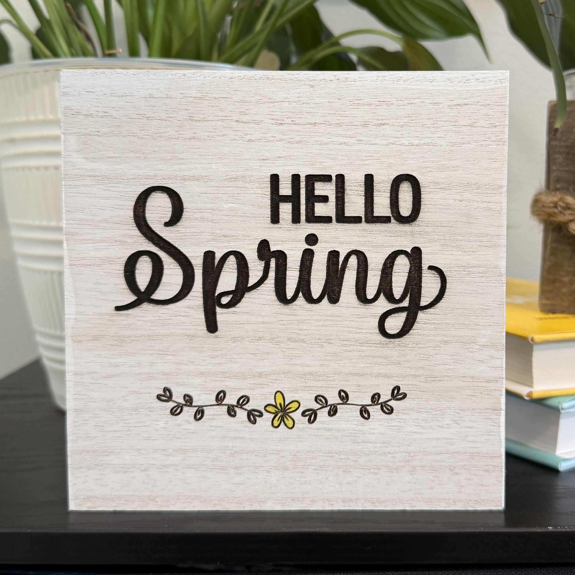 Hello Spring wood shelf sitter with hand-painted yellow flower accent displayed on a black shelf with white planter and greenery in background