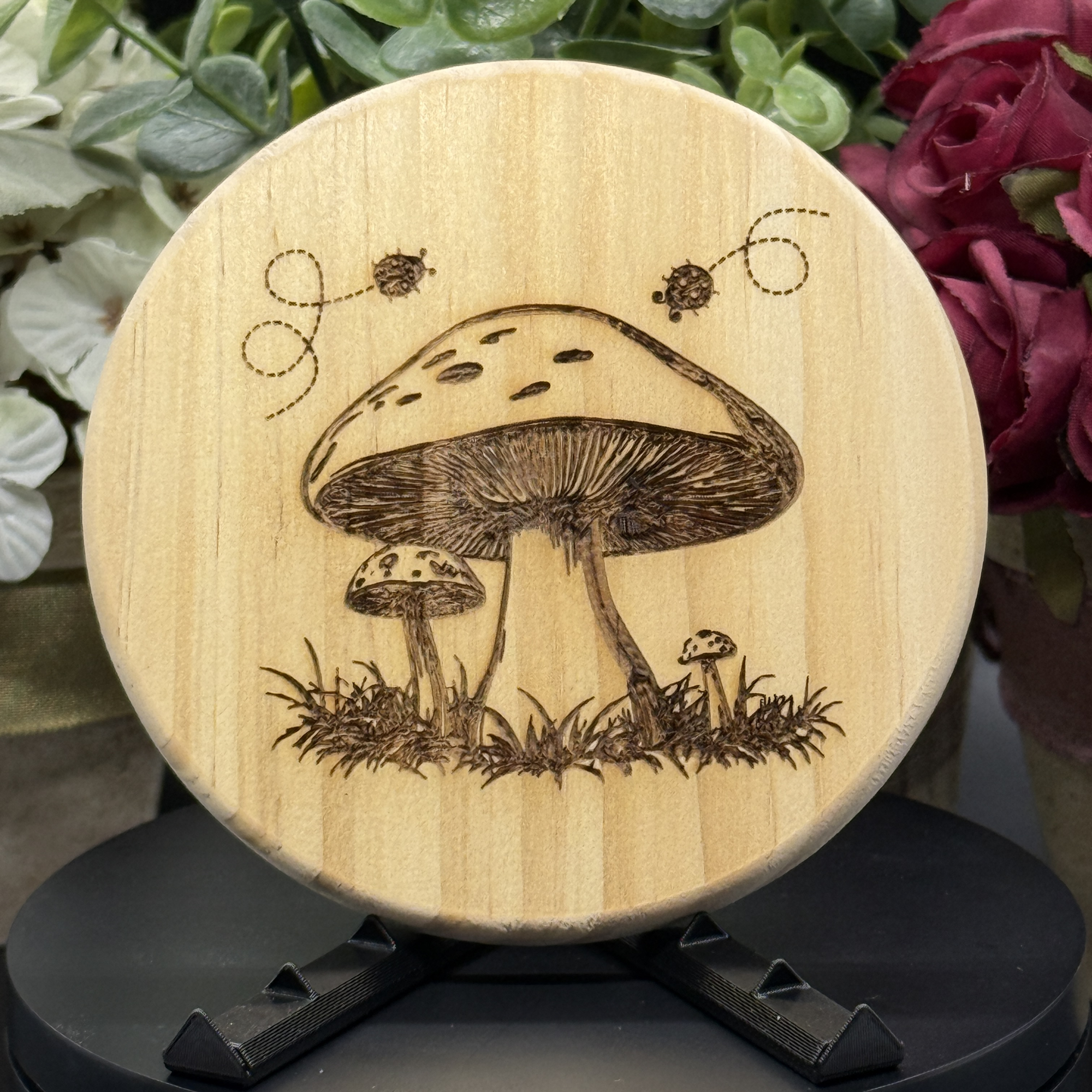 Round pine wood coaster with engraved woodland mushroom scene and flying ladybugs above grass