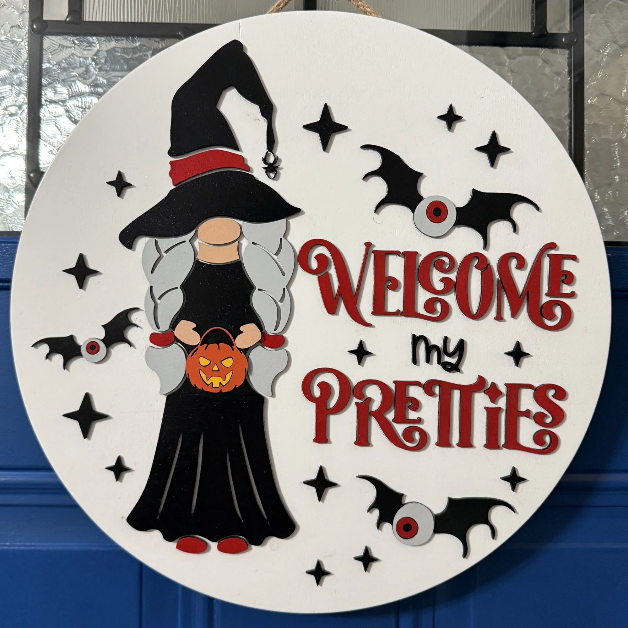 14-inch layered wood “Welcome My Pretties” Halloween door hanger with witch, bats, and stars on a white background, hanging on a blue front door.