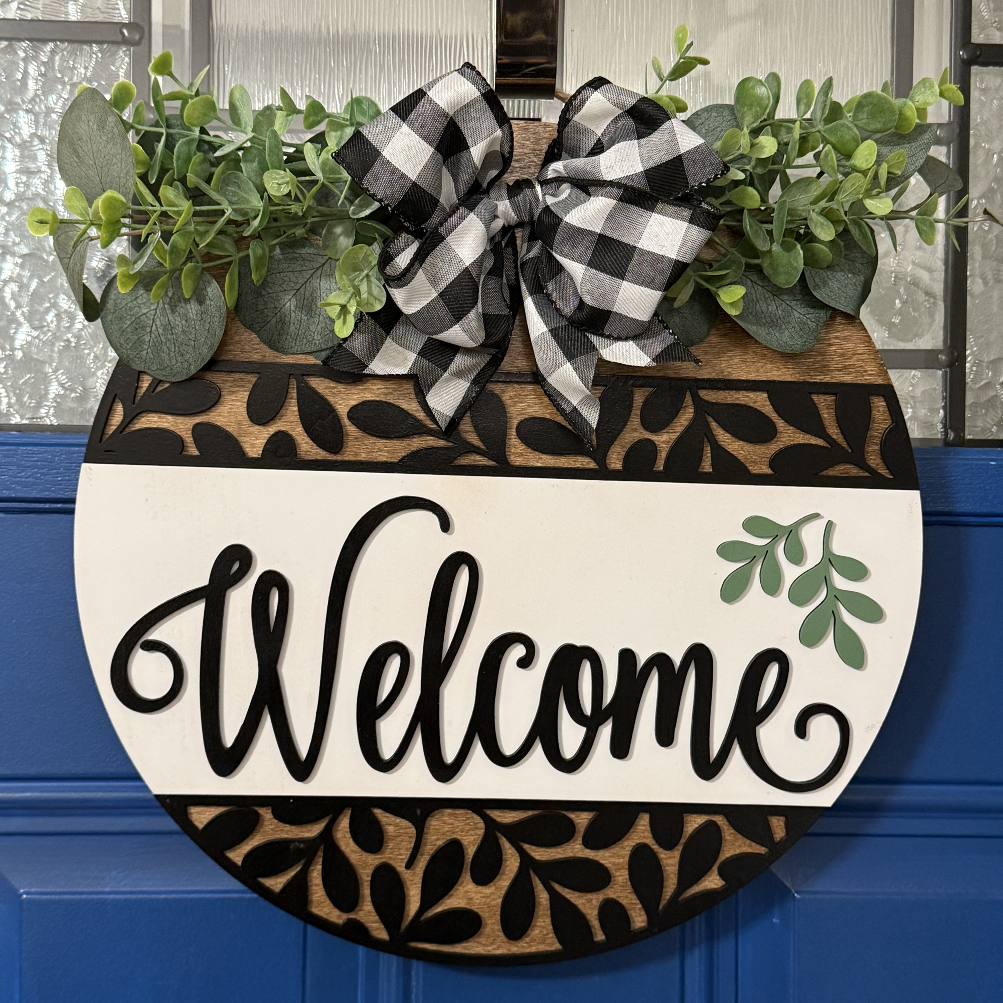 Front view of round buffalo plaid welcome door hanger with greenery, layered leaf design, and white center panel