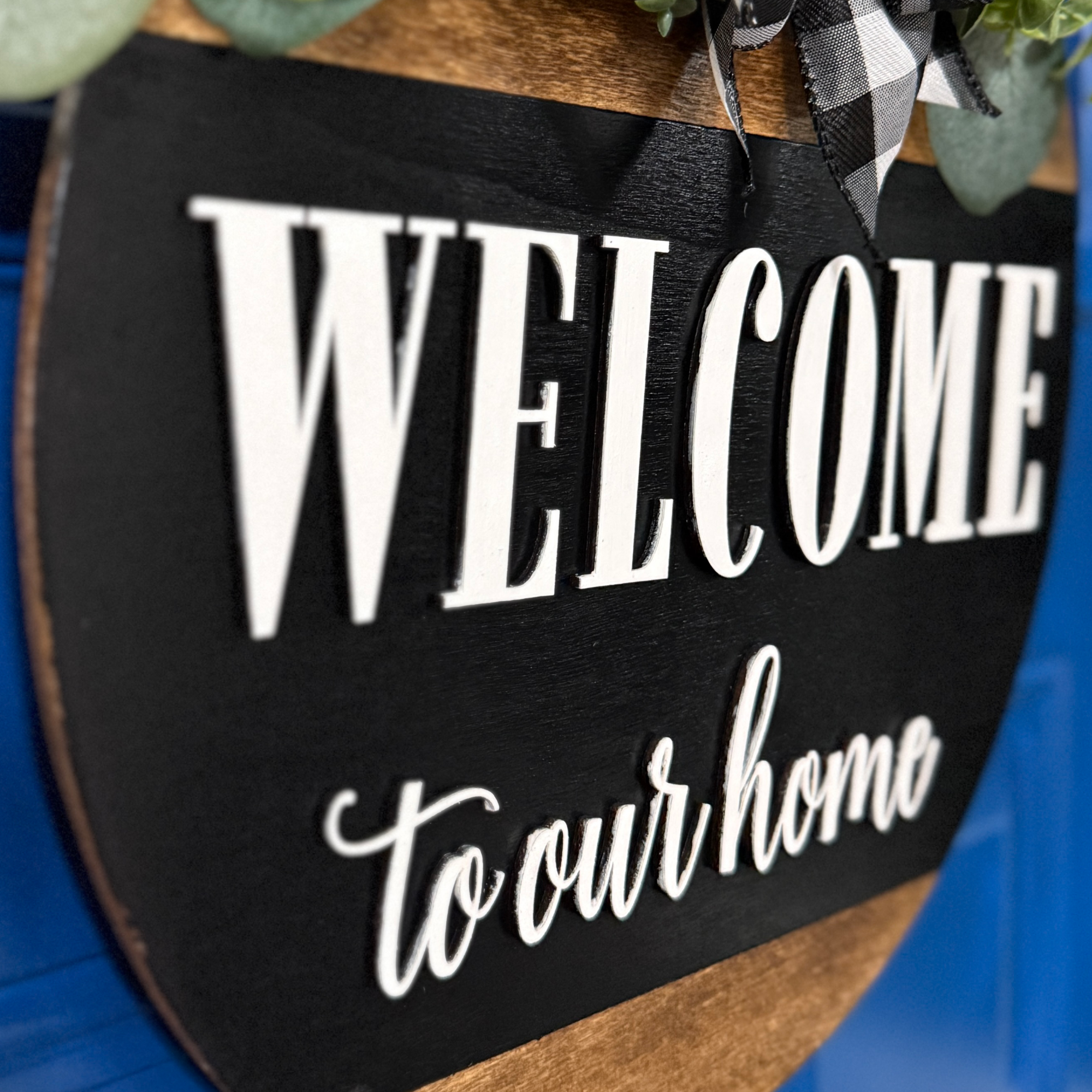 Side angle view of black welcome to our home door hanger with layered wood detail and decorative bow