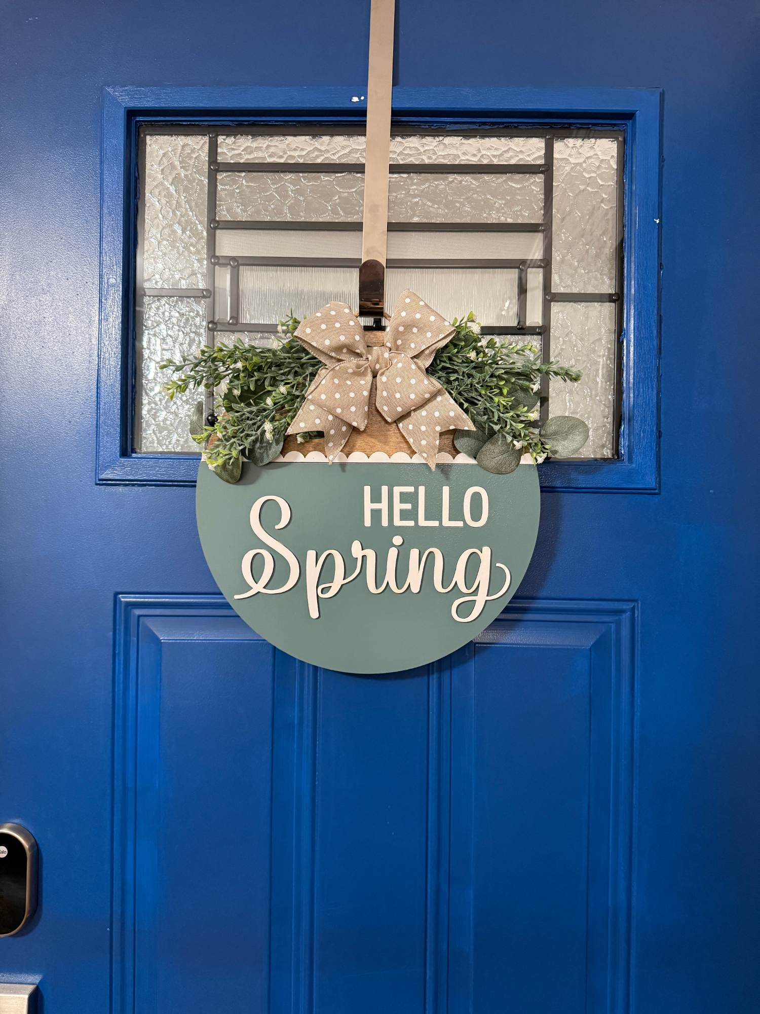 Full view of Hello Spring round wood door hanger displayed on a bright blue front door with neutral bow and greenery accent