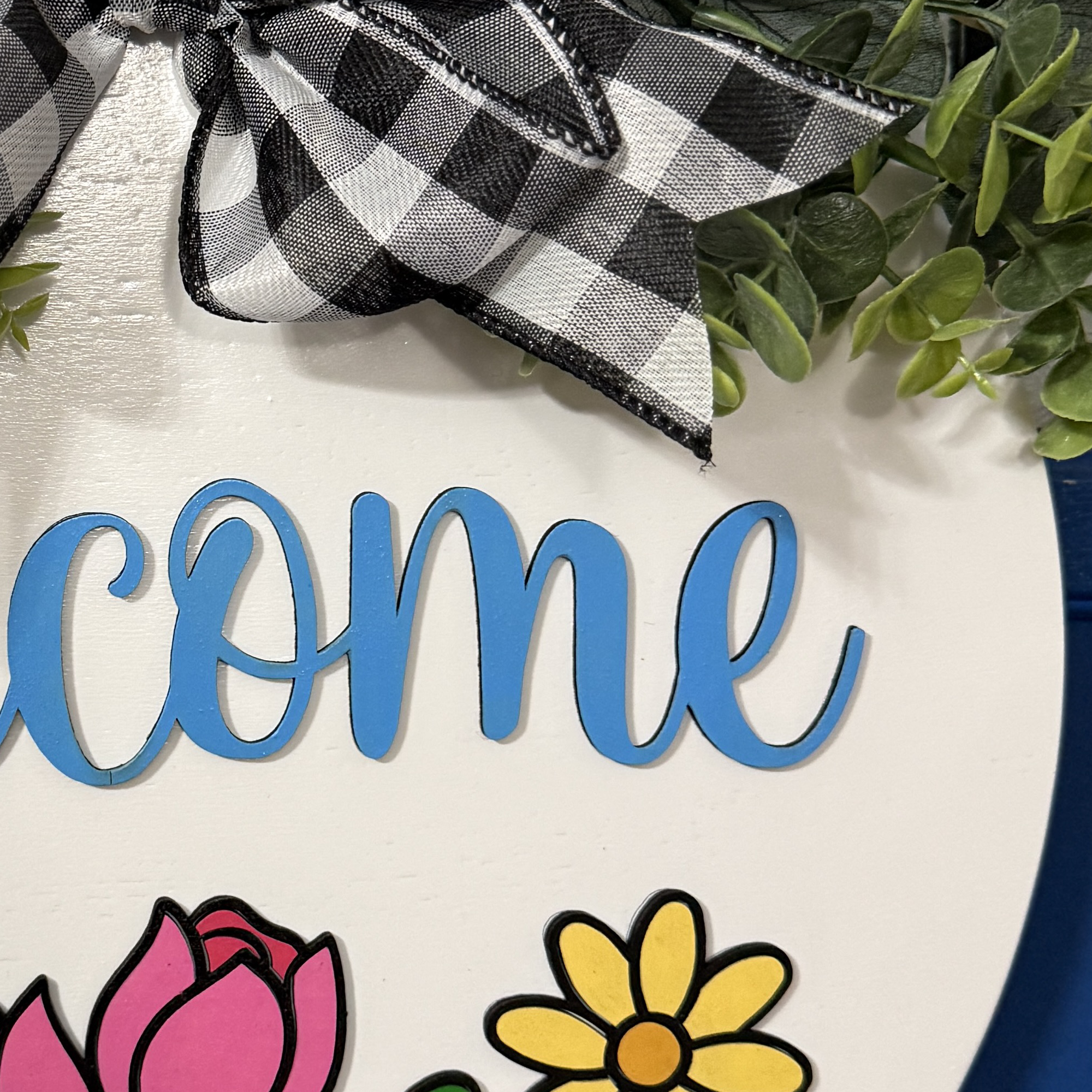 Close-up of raised blue “welcome” script lettering with buffalo check bow and greenery accent