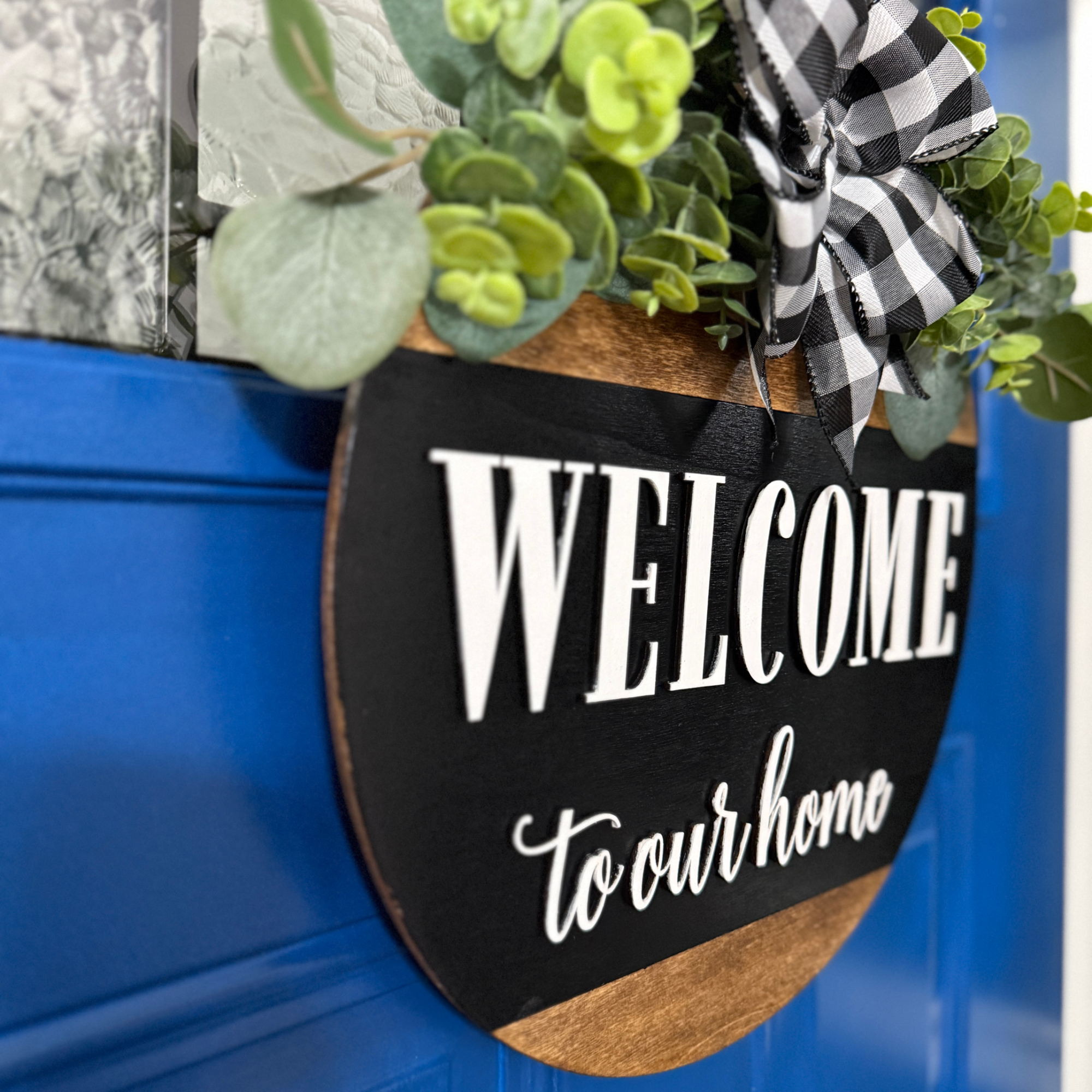 Angled view of welcome to our home door hanger with black painted wood, greenery, and buffalo plaid ribbon bow