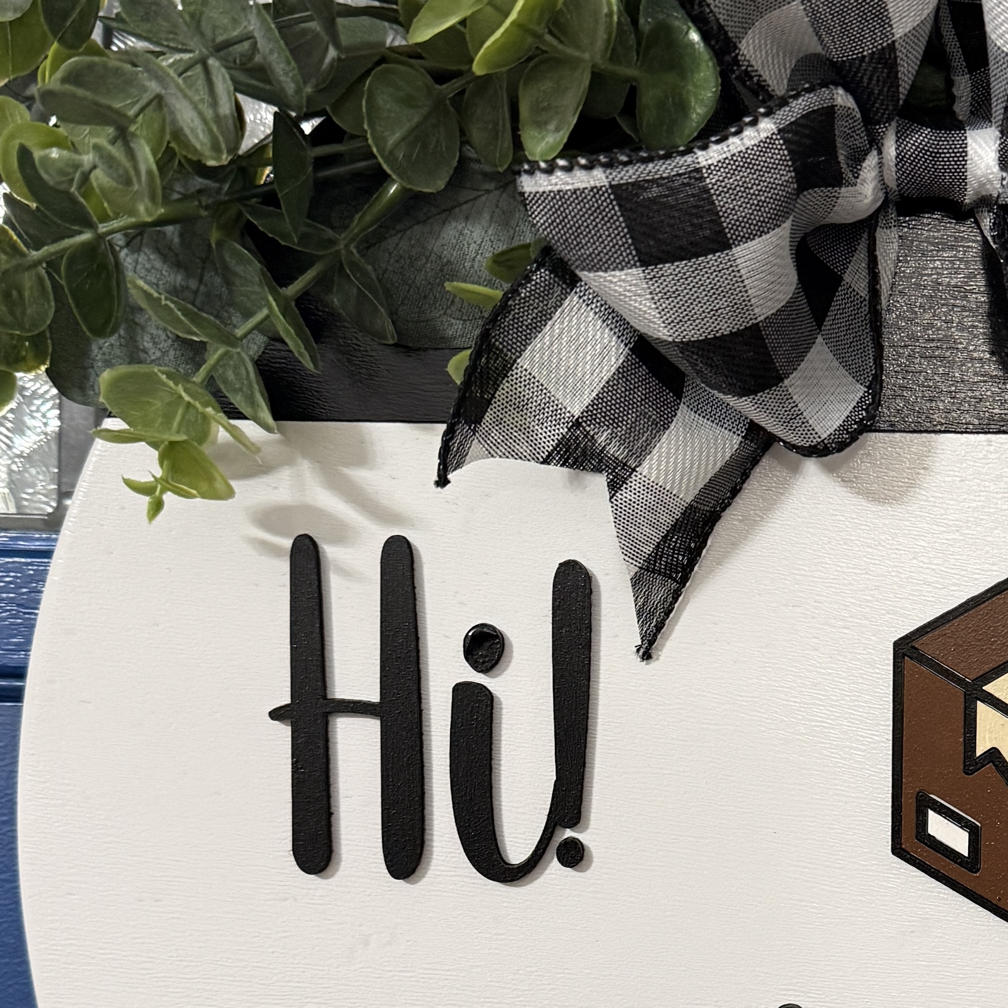Close-up of top portion of white wooden door hanger showing raised “Hi!” lettering, layered package graphic, greenery, and black and white buffalo plaid bow.