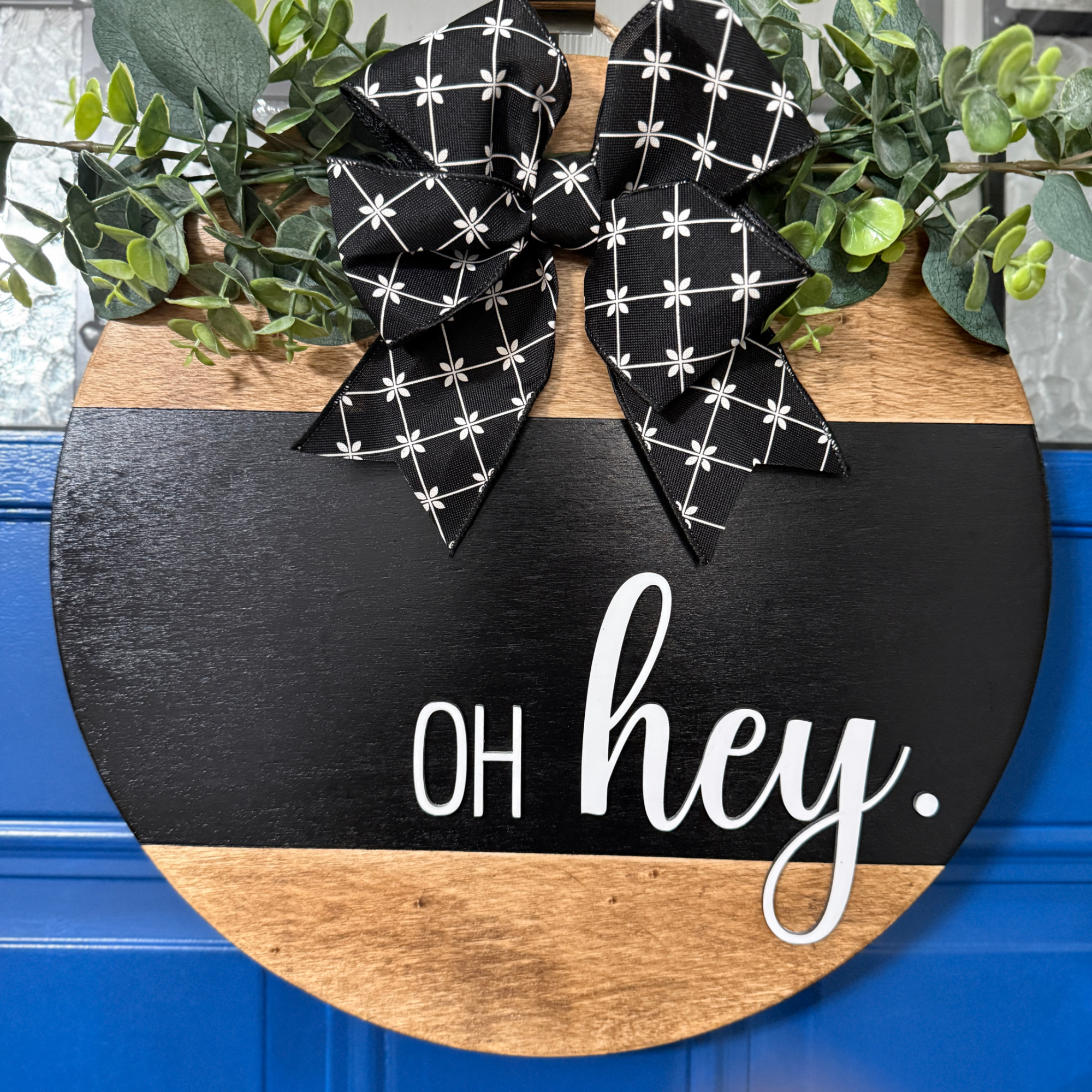 Black and natural wood round oh hey door hanger with white lettering, greenery accents, and black patterned bow on blue front door