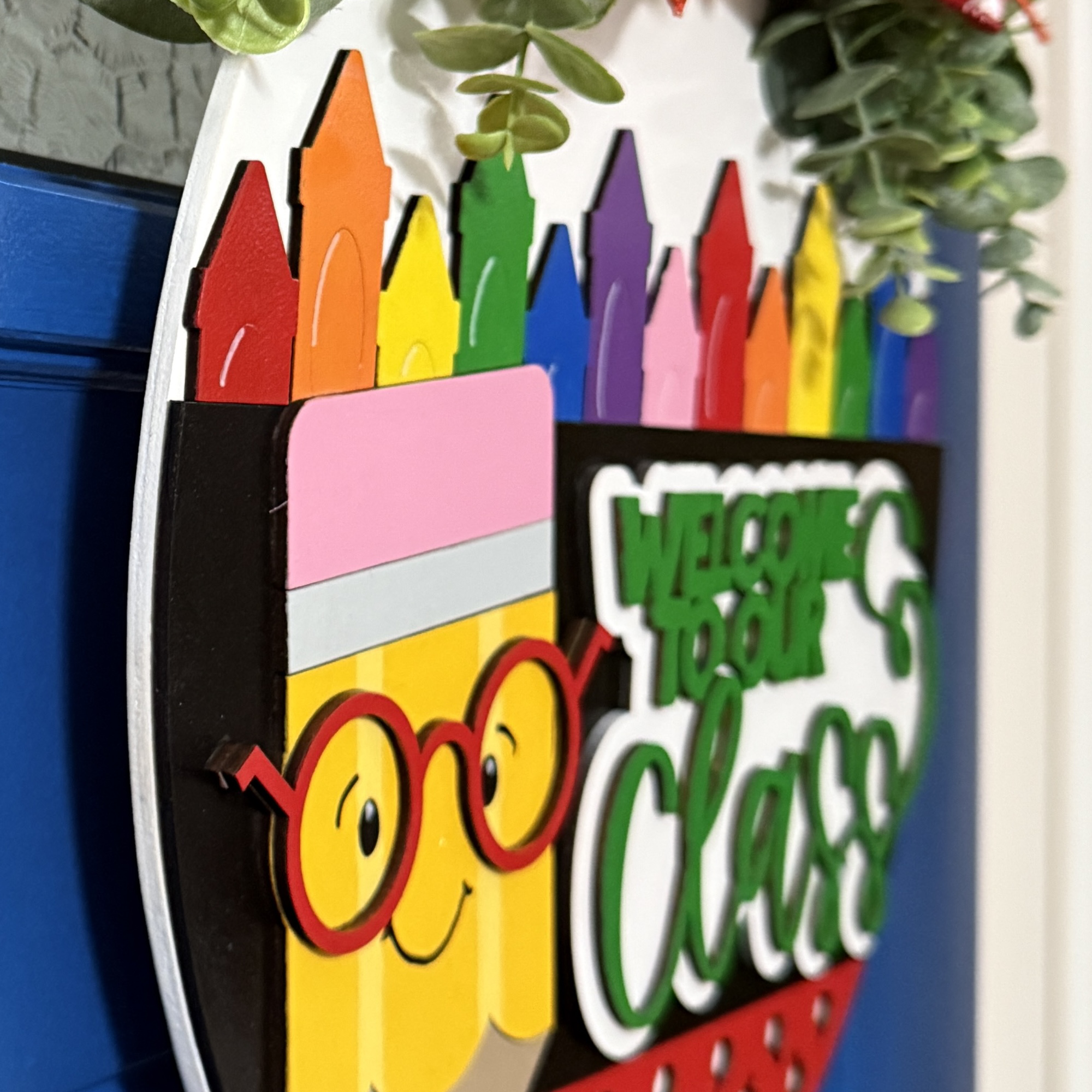 Side view of “Welcome to Our Class” door hanger featuring smiling pencil, rainbow crayon row, and red/yellow polka dot bow with greenery on a dark blue door.